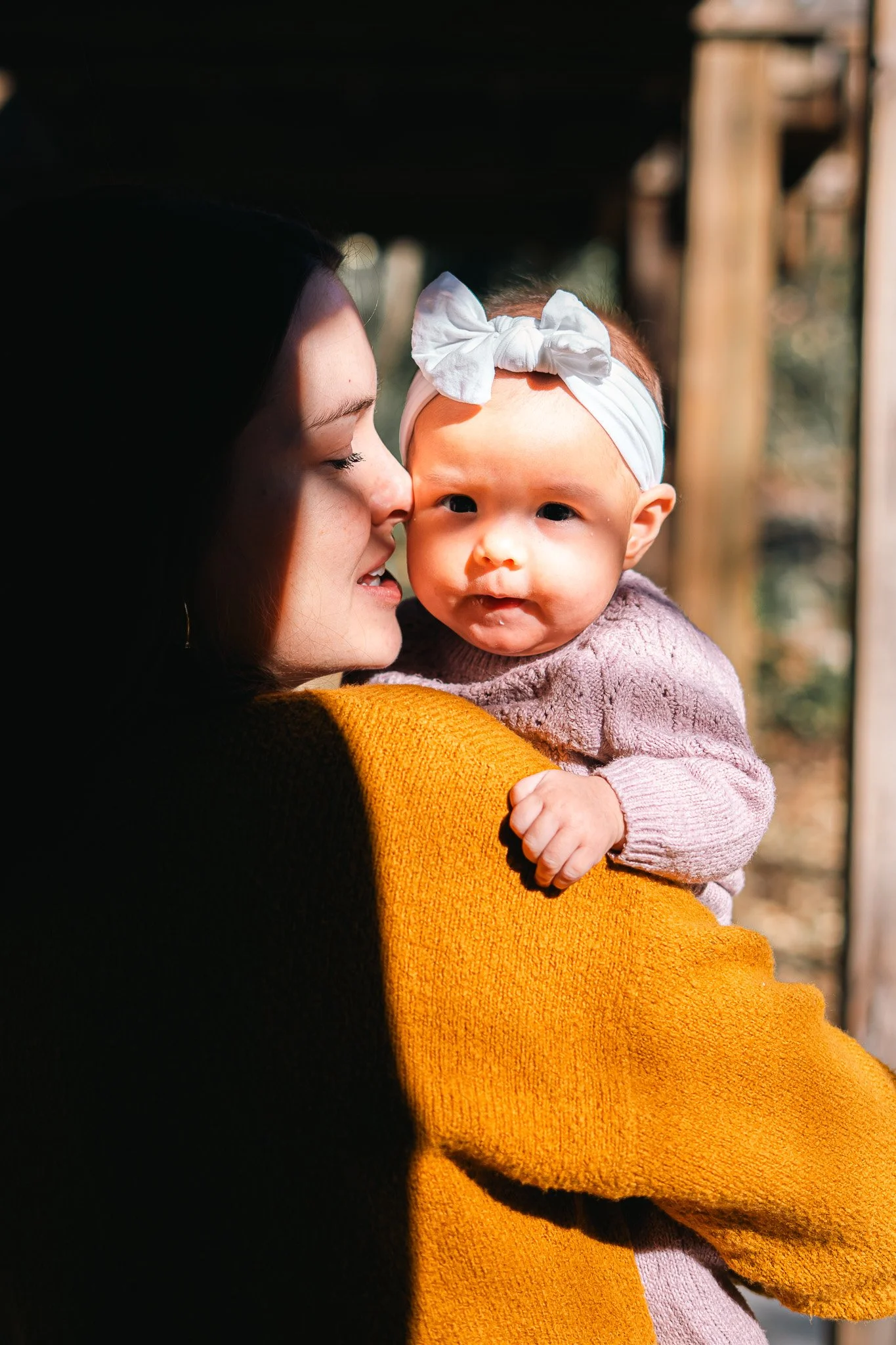 A woman holding a baby girl outdoors, with the woman whispering or nuzzling close to the baby. The baby wears a white headband with a bow and a light purple sweater, while the woman wears a mustard yellow sweater. Sunlight highlights their faces.