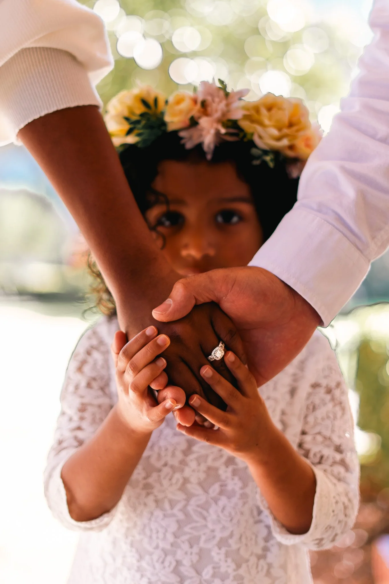 A young girl with a flower crown looks intensely at the camera while holding hands with two adults, one wearing a white shirt and the other in a white sleeve. The focus is on the hands, with the girl in the background.