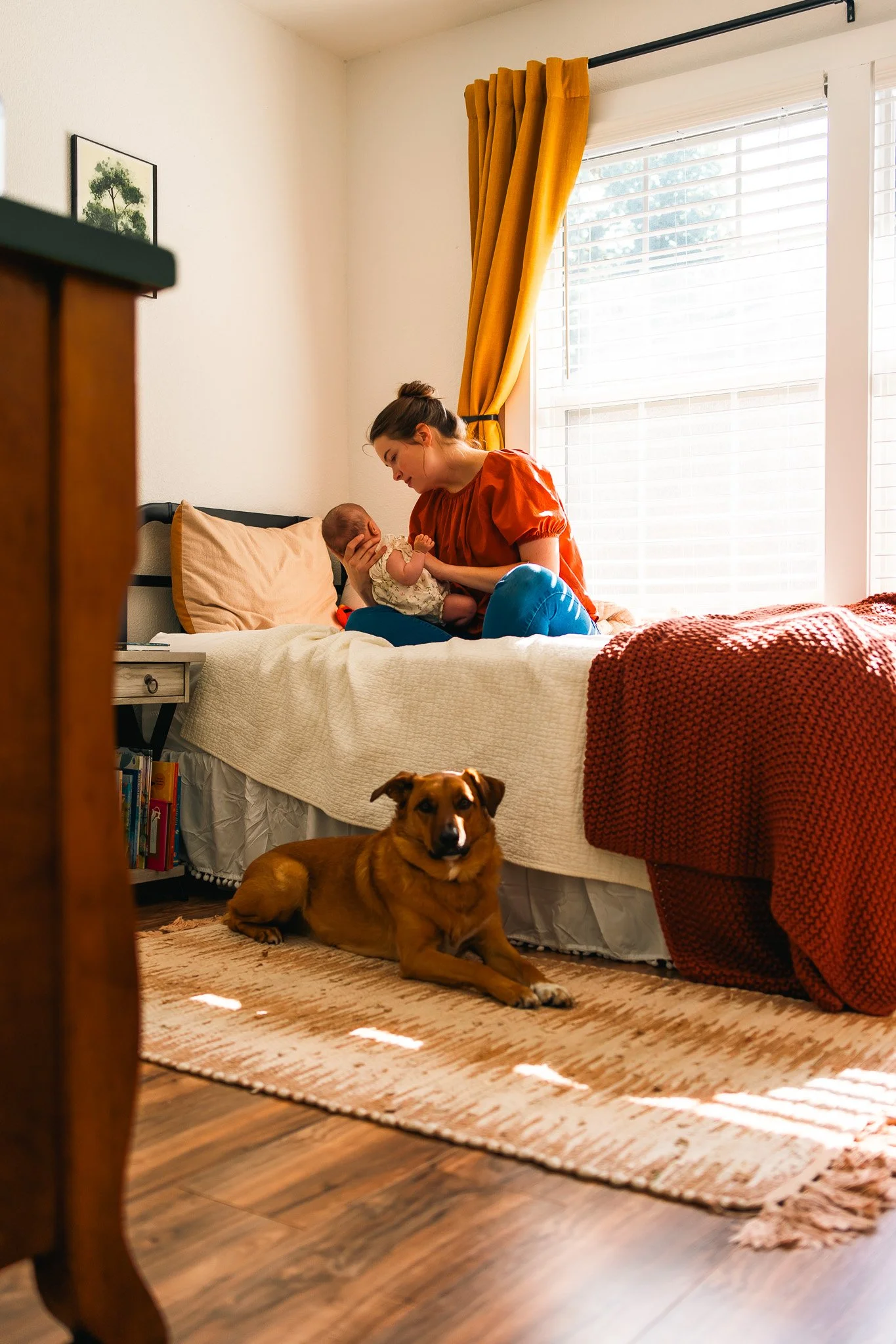 A woman holding a baby on a bed in a sunlit bedroom with a brown dog lying on a rug nearby.