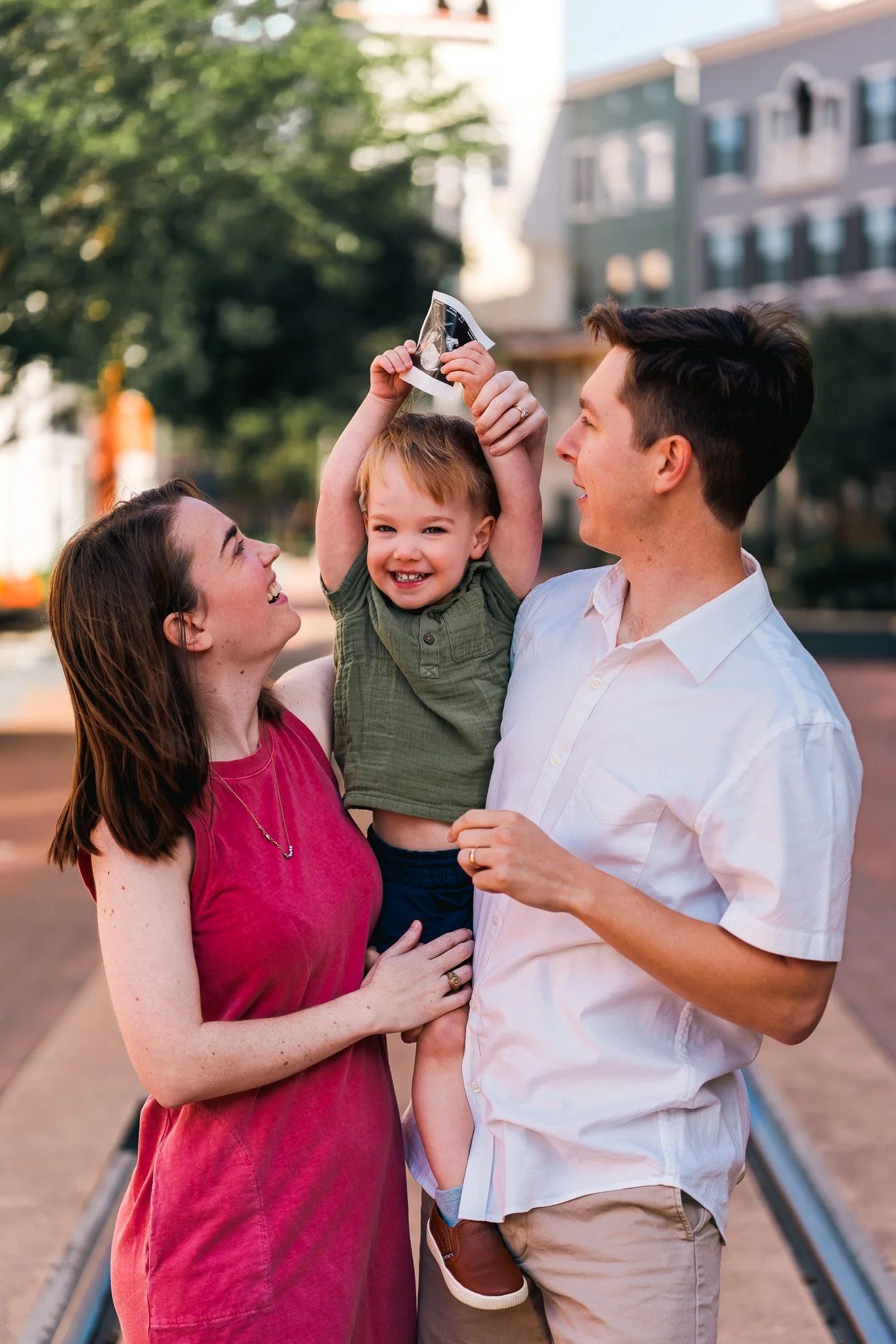 A family of three enjoying a moment outdoors, with a woman, man, and young boy. The boy, holding a small ultrasound image, is lifted by the woman and looks happy, while the man smiles at him. They are near a street with trees and buildings in the bac