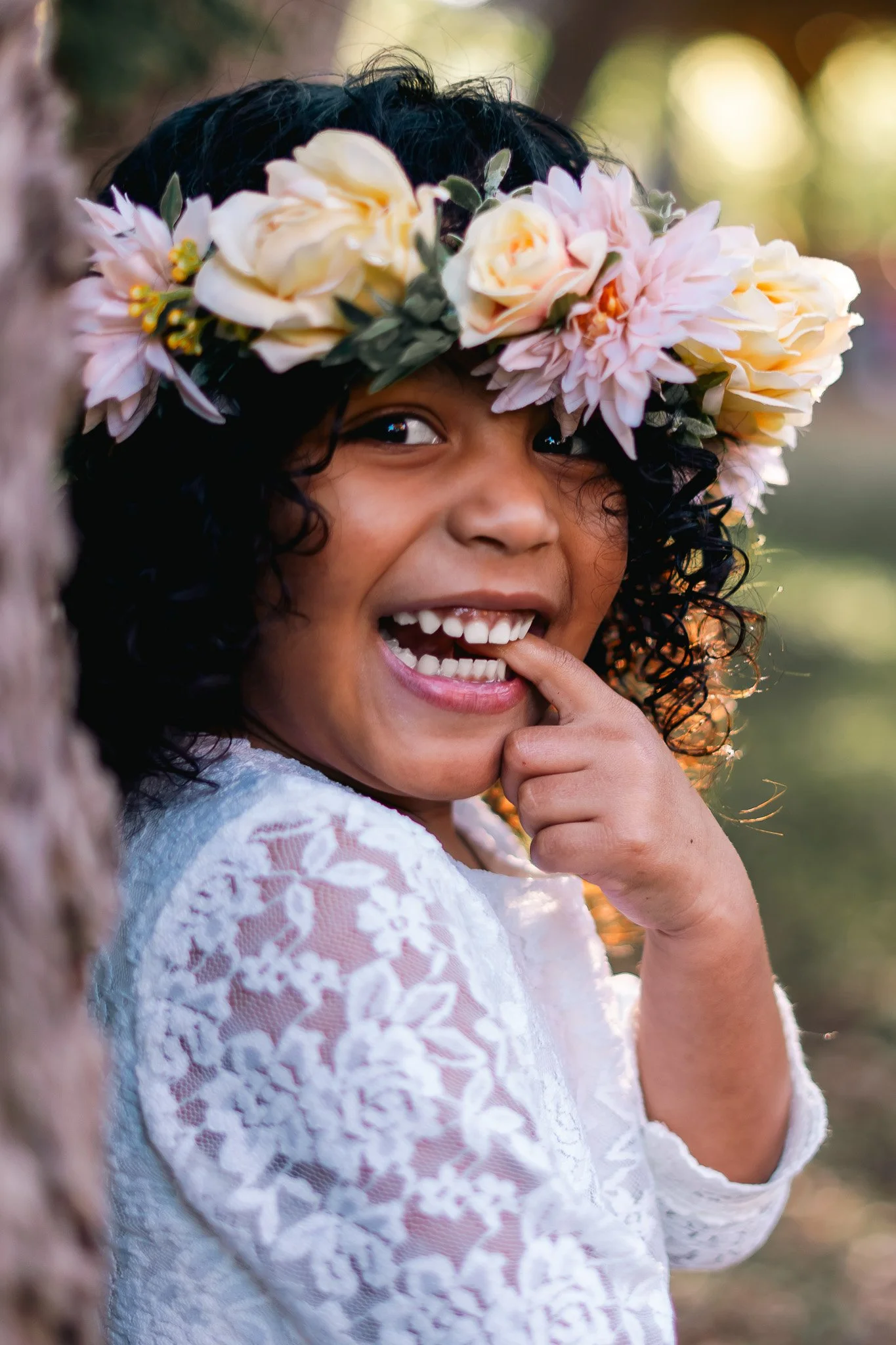 A young girl wearing a floral crown, smiling, with one hand near her mouth, outside near a tree.