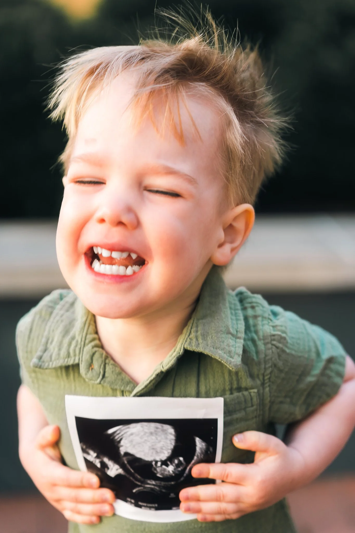 Young boy with tousled blonde hair, smiling with eyes closed, holding an ultrasound picture of a fetus on his chest.