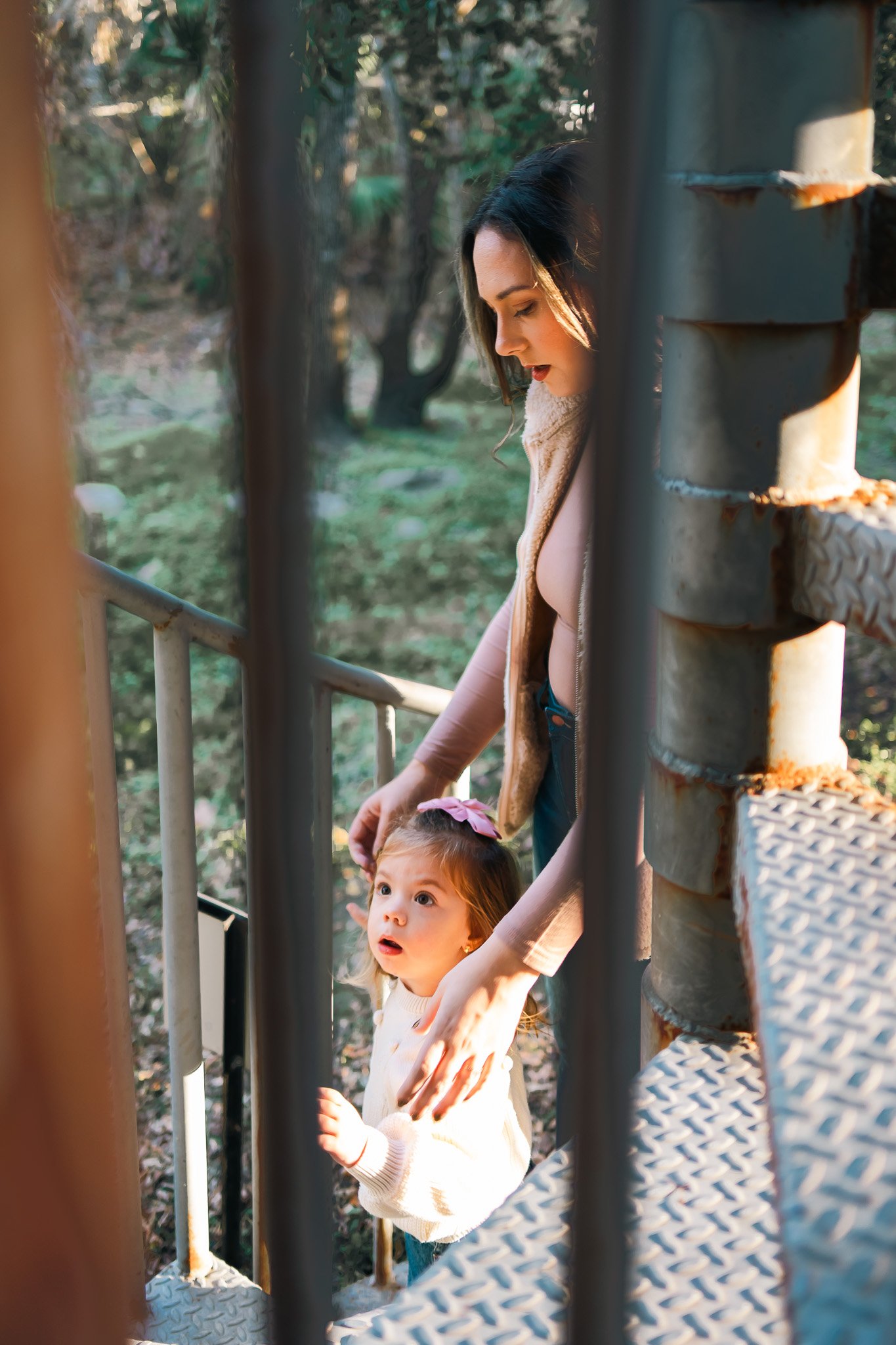 A woman and a young girl on an outdoor metal staircase surrounded by trees. The woman is looking down at the girl, who has an expression of surprise or curiosity. The scene is lit by natural sunlight.