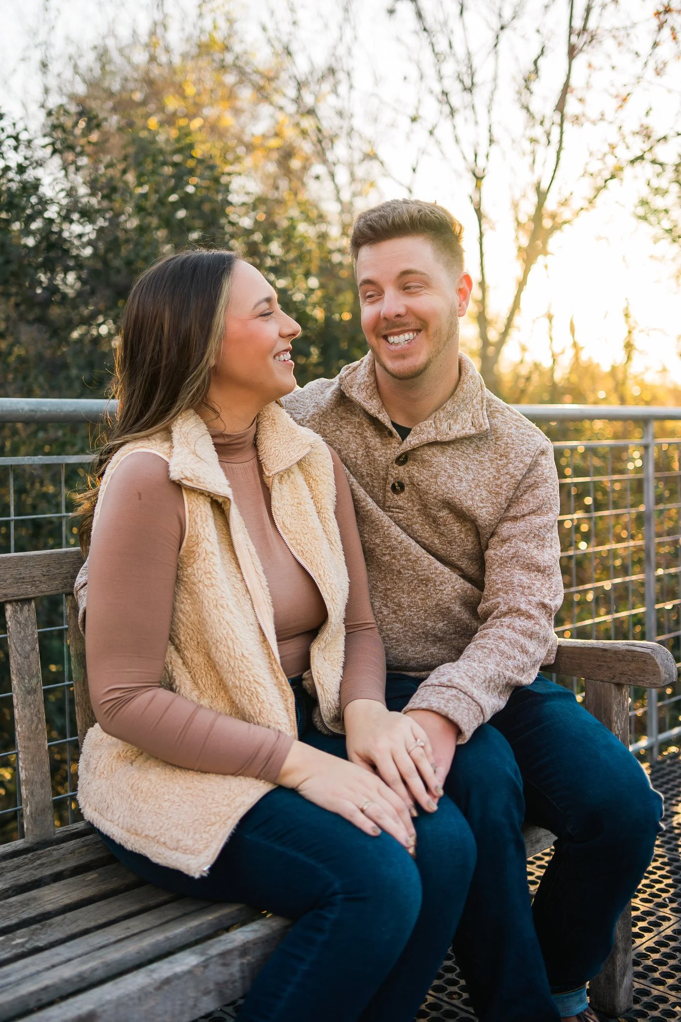 A couple sitting on a wooden bench outdoors, smiling at each other during sunset with trees in the background.