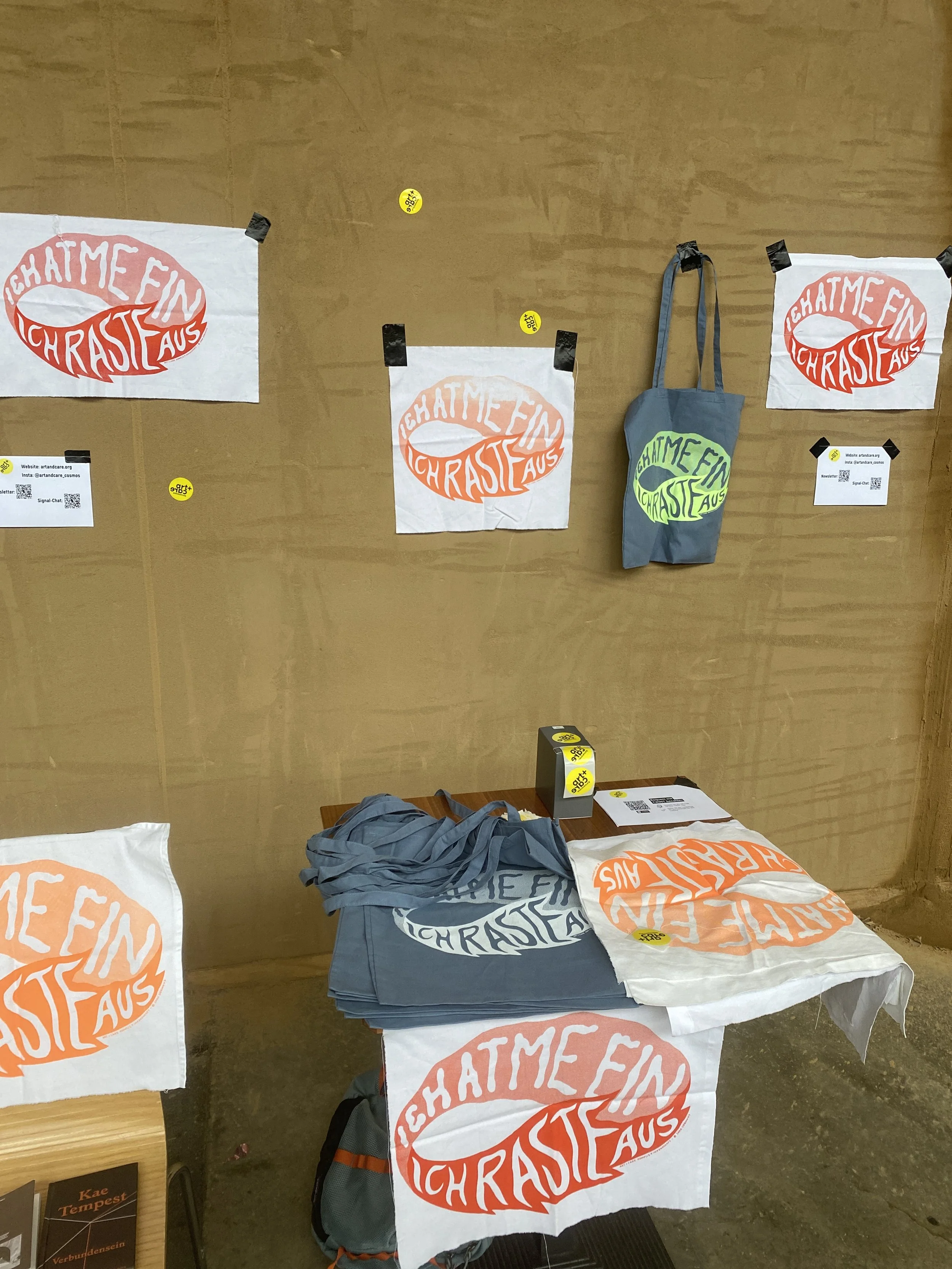 Verschiedene T-Shirts mit dem Aufdruck 'Chatme Ein Chraste Aus' in rot und orange, sowie Einkaufstaschen mit demselben Aufdruck, auf einem Tisch und an einer Wand befestigt.