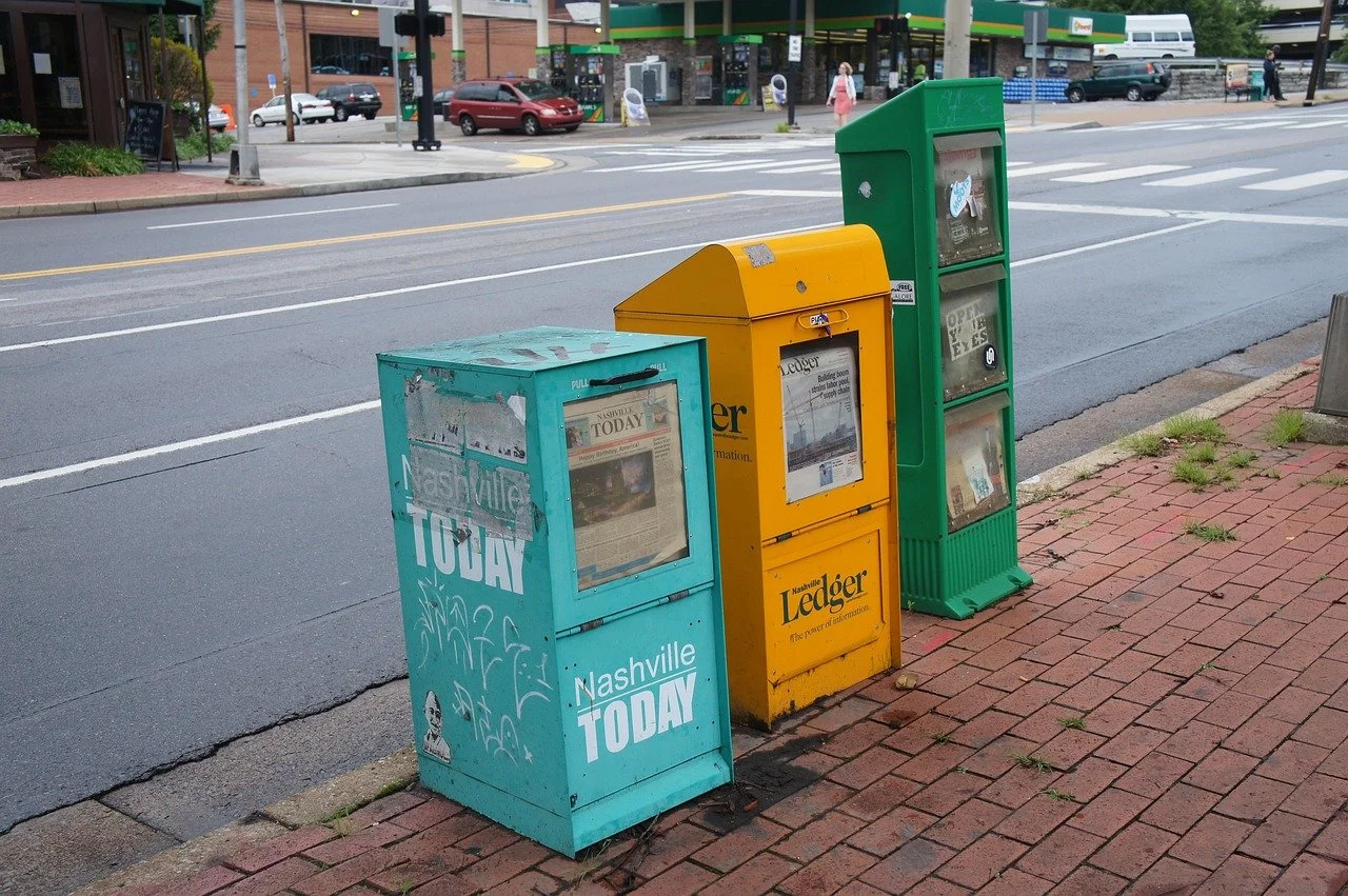 Three newspaper vending machines in different colors on a brick sidewalk next to a street, with a storefront and cars visible in the background.