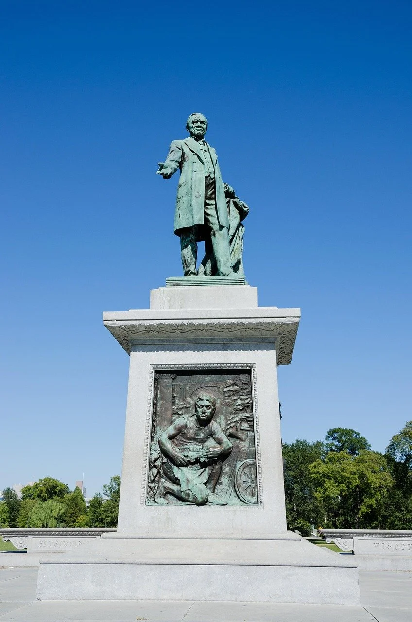 Statue of a man in a coat with a child behind him and relief carvings at the base, set against a clear blue sky and green trees.