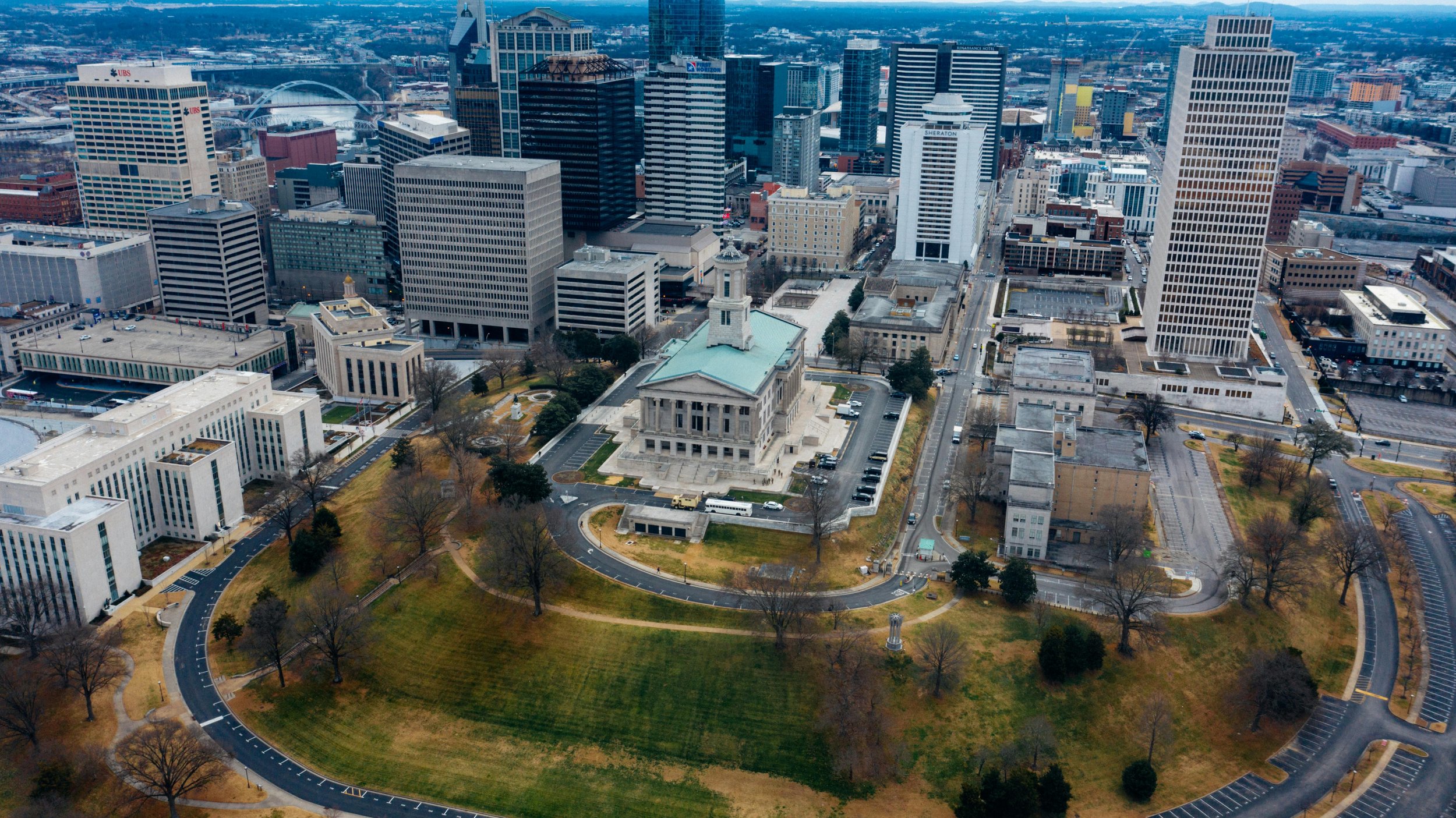 Aerial view of Nashville, Tennessee, featuring the Tennessee State Capitol in the city center surrounded by skyscrapers and a park with walking paths and trees.