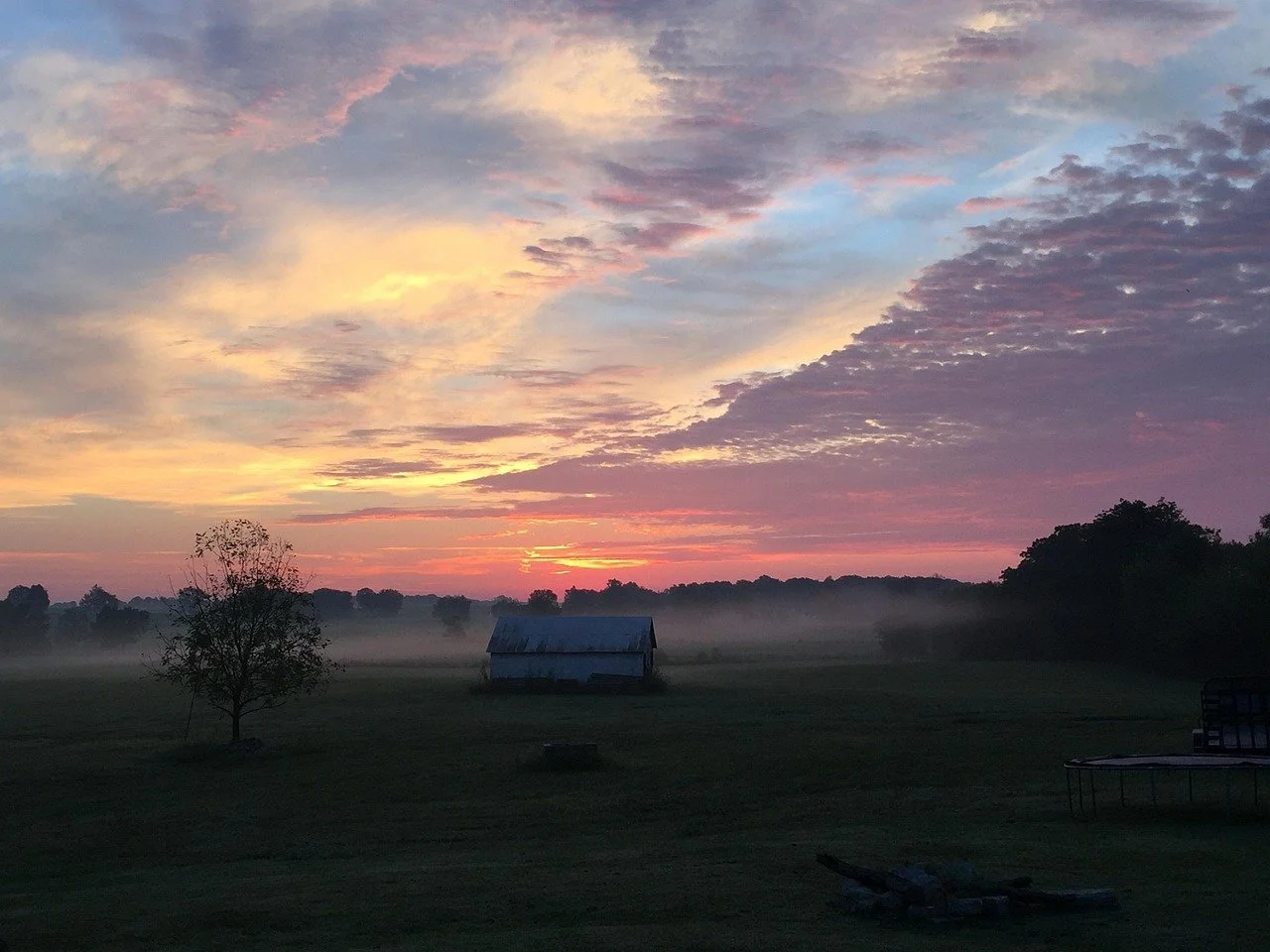 Sunrise over a rural landscape with a small barn, a tree, mist on the ground, and a trampoline on the right, under a colorful sky with clouds.