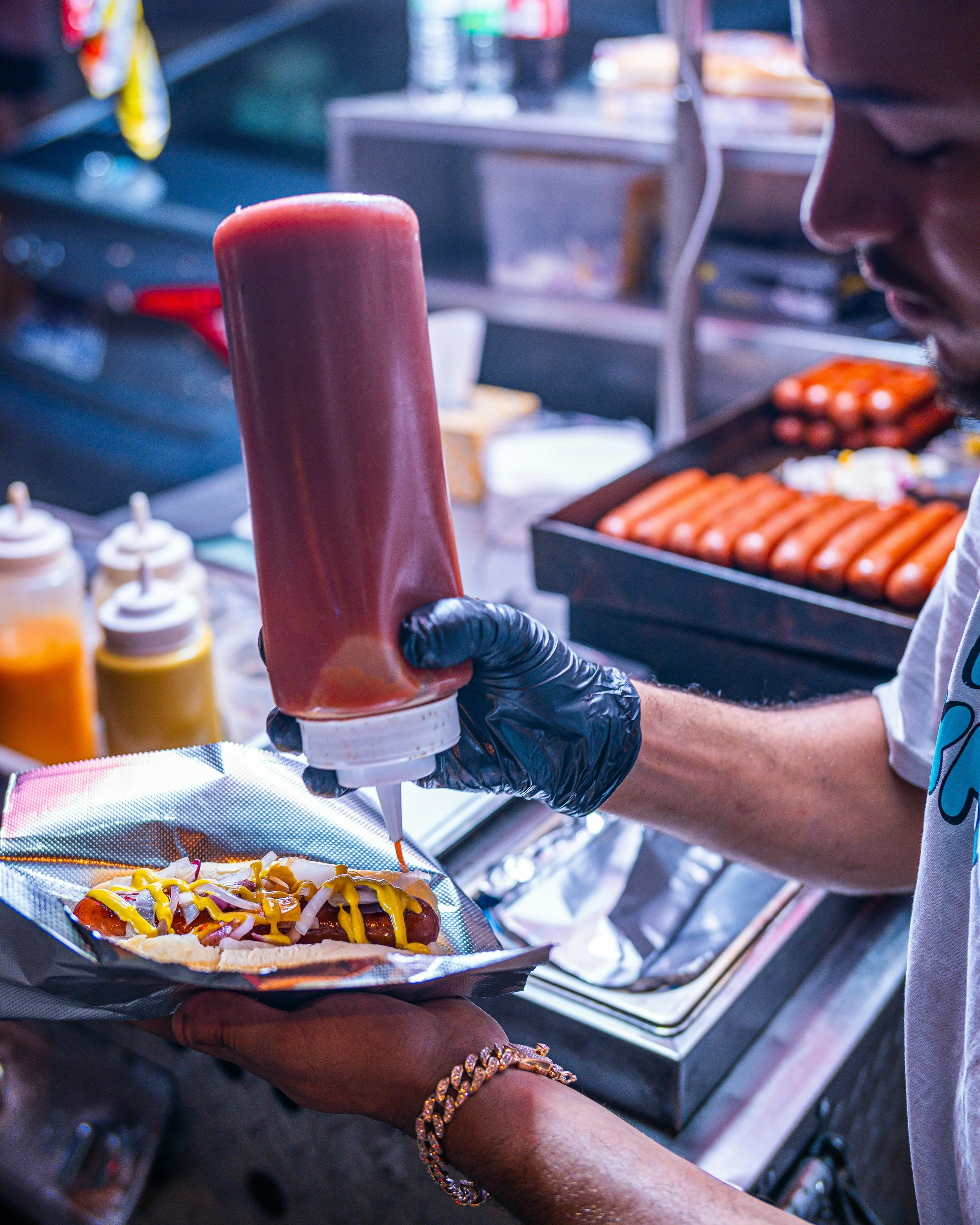 Person wearing a gold bracelet and black gloves preparing a hot dog with mustard, ketchup, and onions at a food vendor stand, with condiments and hot dogs visible in the background.