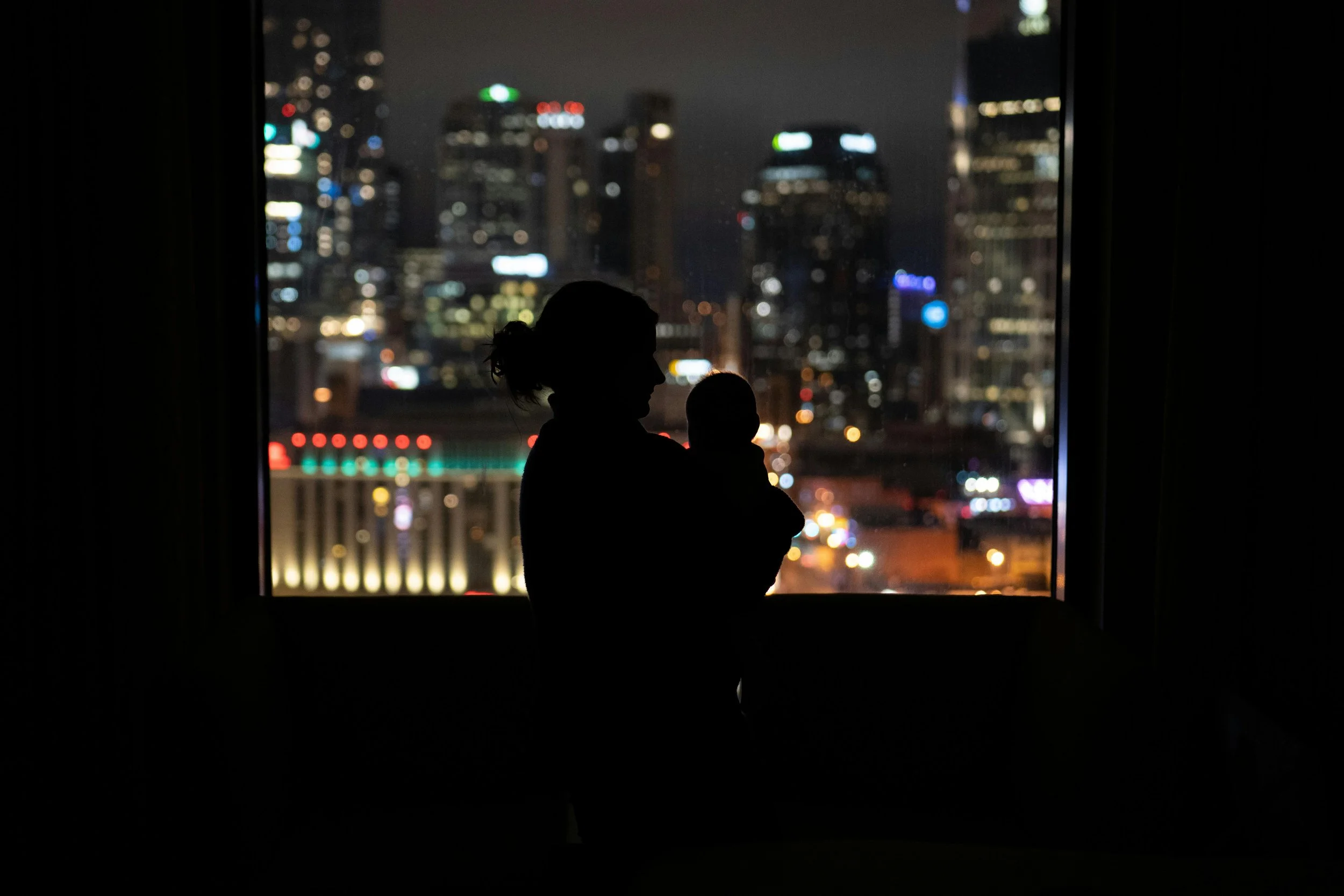 Silhouette of a woman holding a baby, standing in front of a large window overlooking a brightly lit city skyline at night.