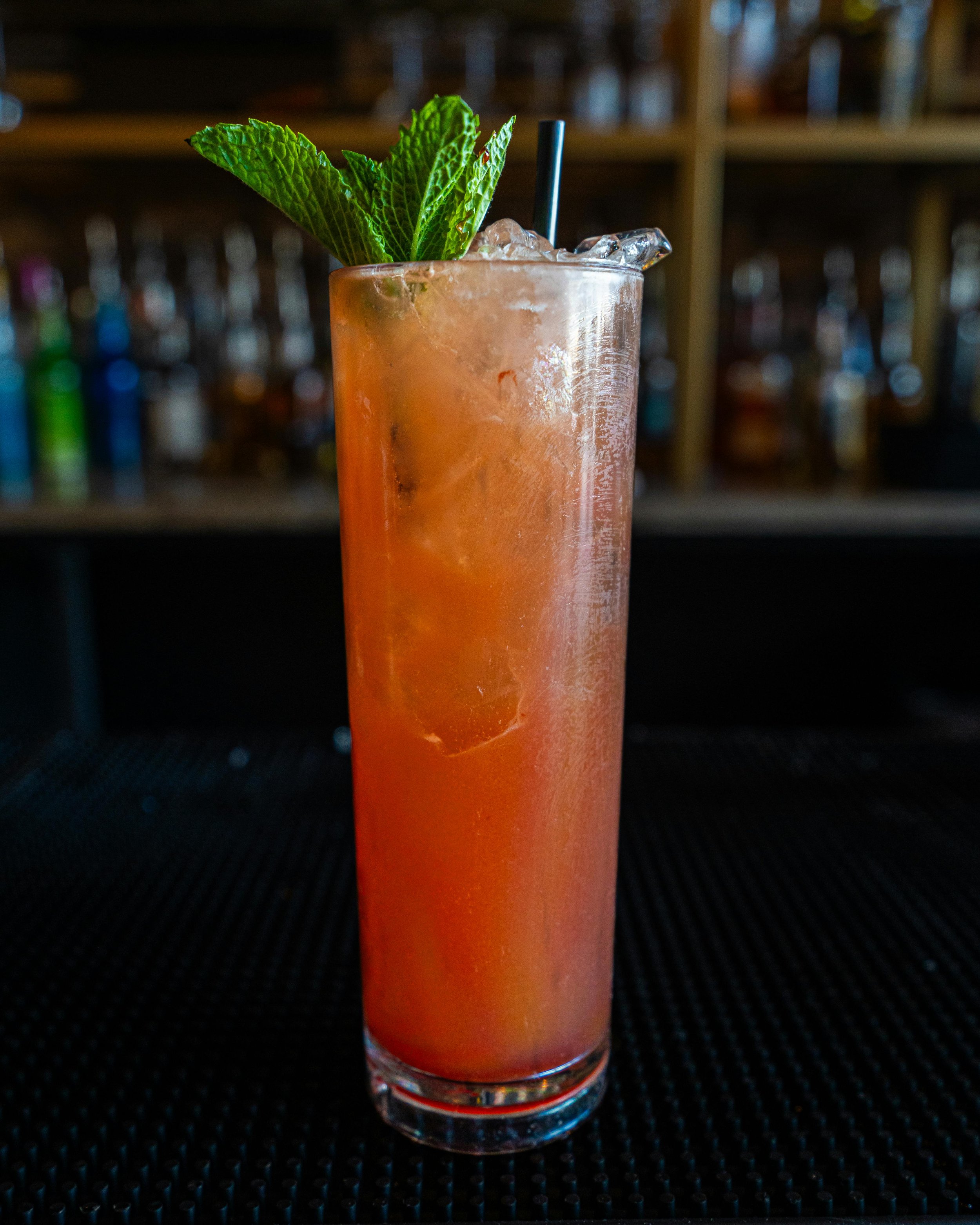 Tall glass cocktail with orange-colored drink, ice cubes, garnished with mint leaves and a black straw, on a black surface bar backdrop.