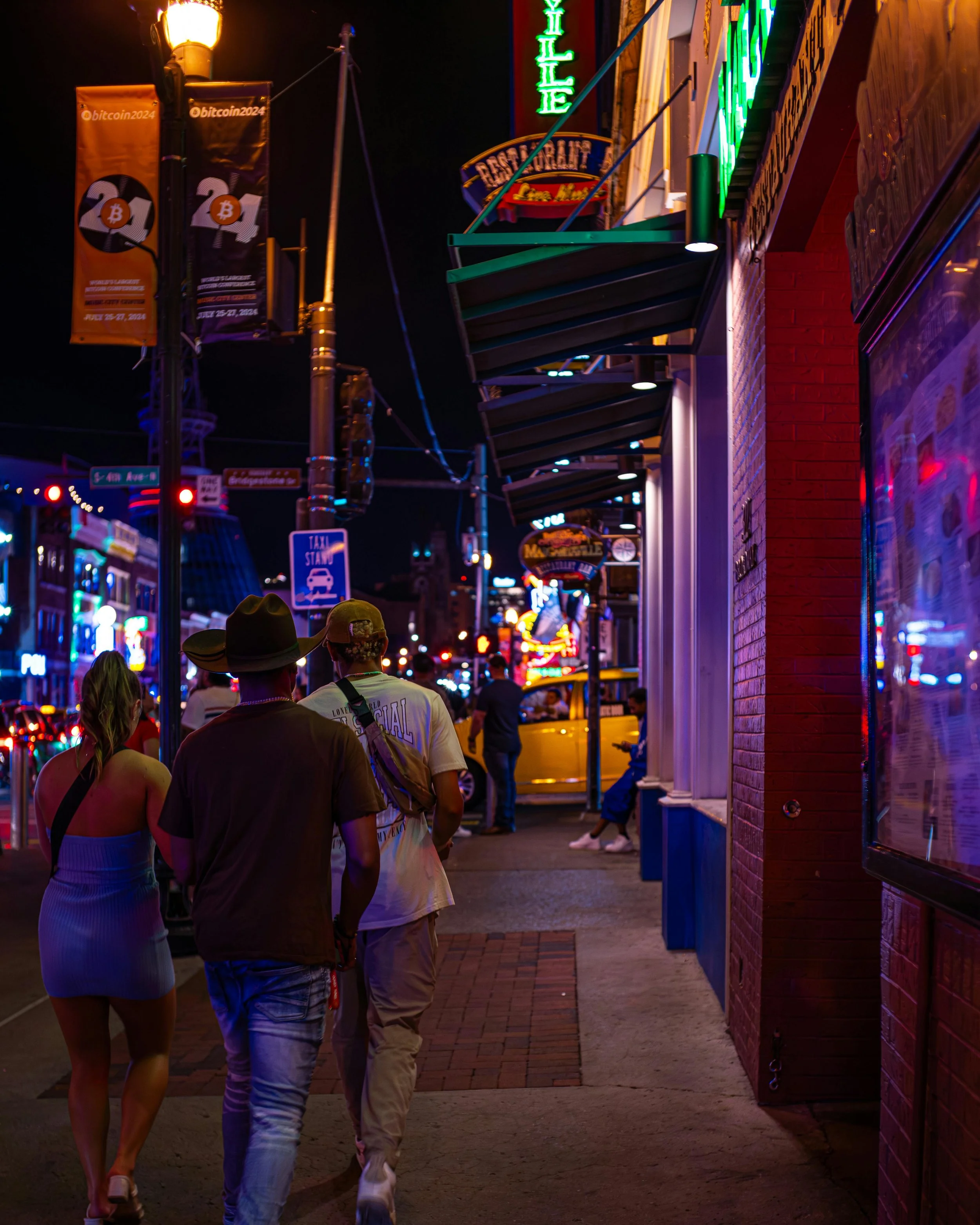 People walking on a brightly lit city street at night with colorful neon signs and illuminated storefronts.