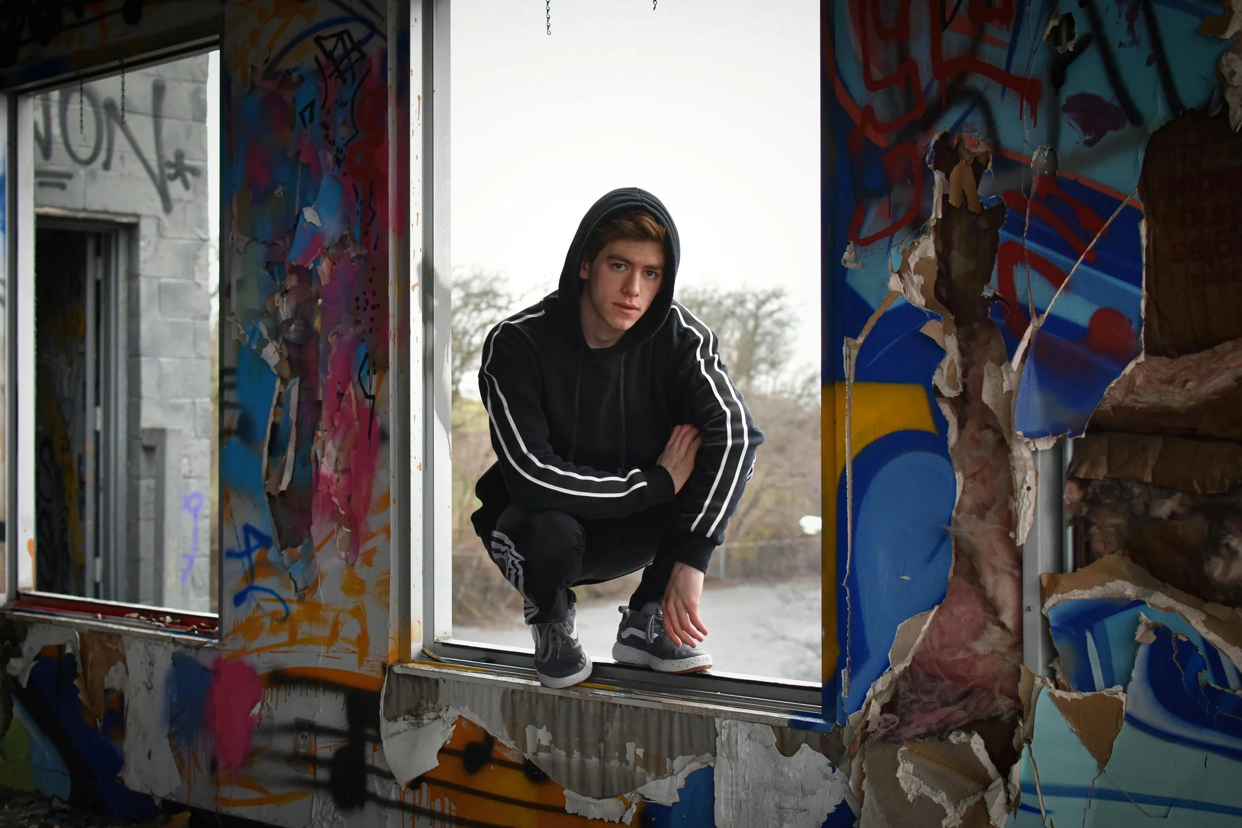 A young man wearing a black hoodie and black Adidas sweatpants crouches on the window sill of a partially destroyed building with graffiti-covered walls.