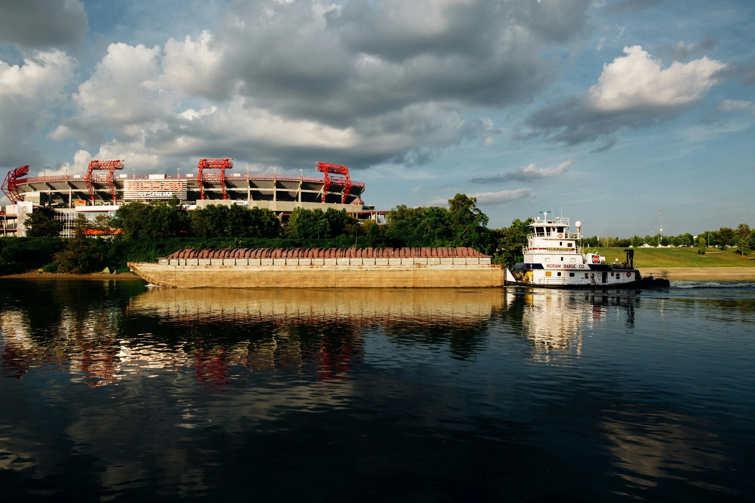 A barge being pushed by a tugboat on a river, with a stadium and trees in the background under a cloudy sky.