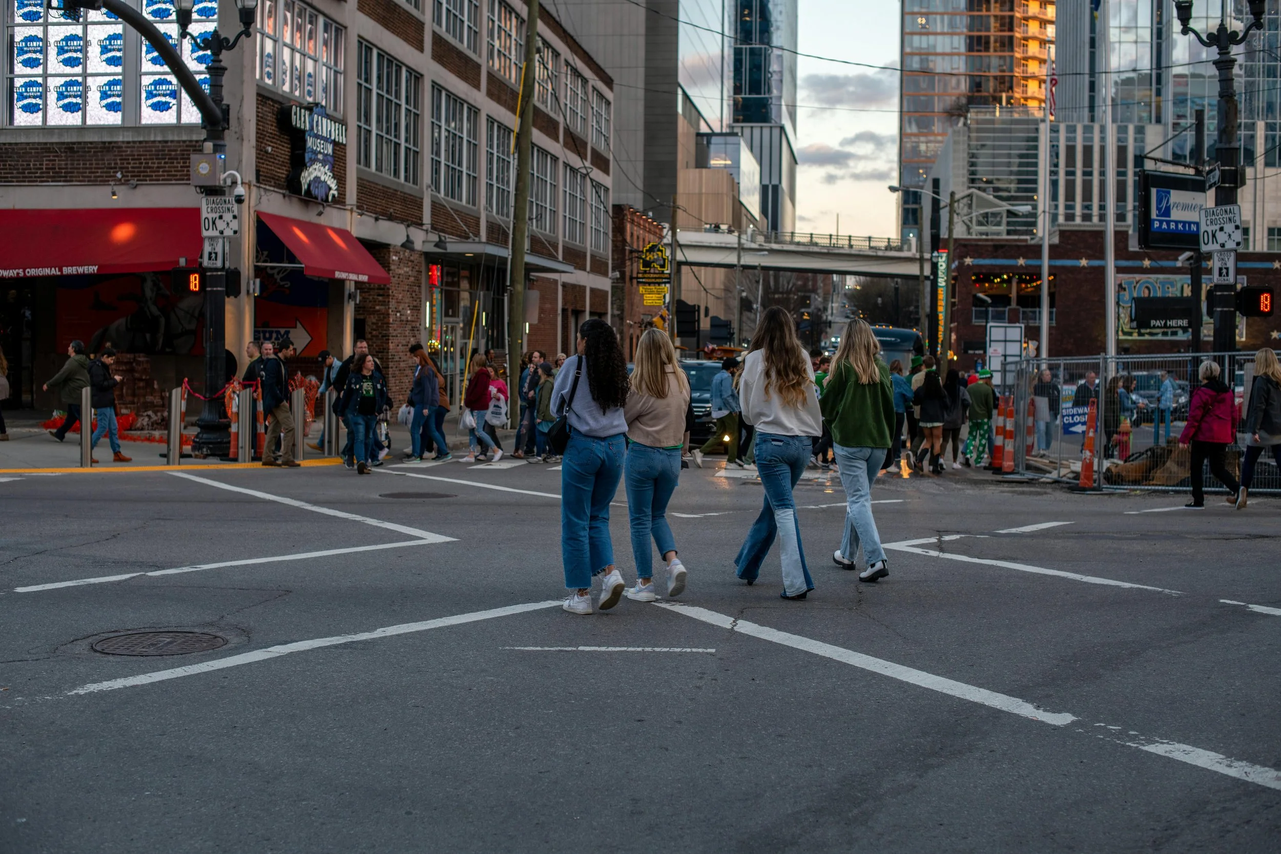 People crossing the street in an urban cityscape at dusk with tall buildings and storefronts in the background.