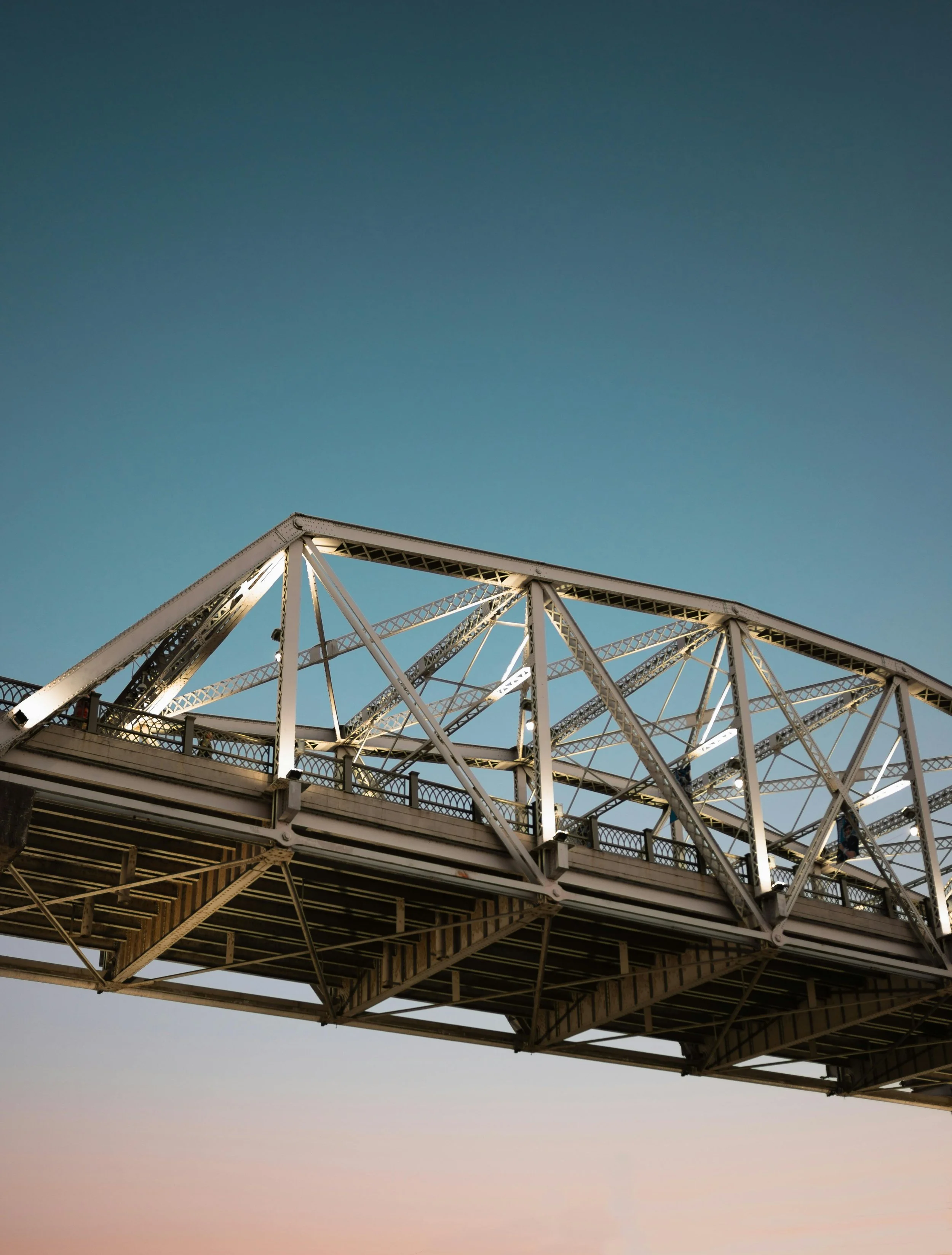 Steel bridge with truss structure over a calm landscape during sunset.