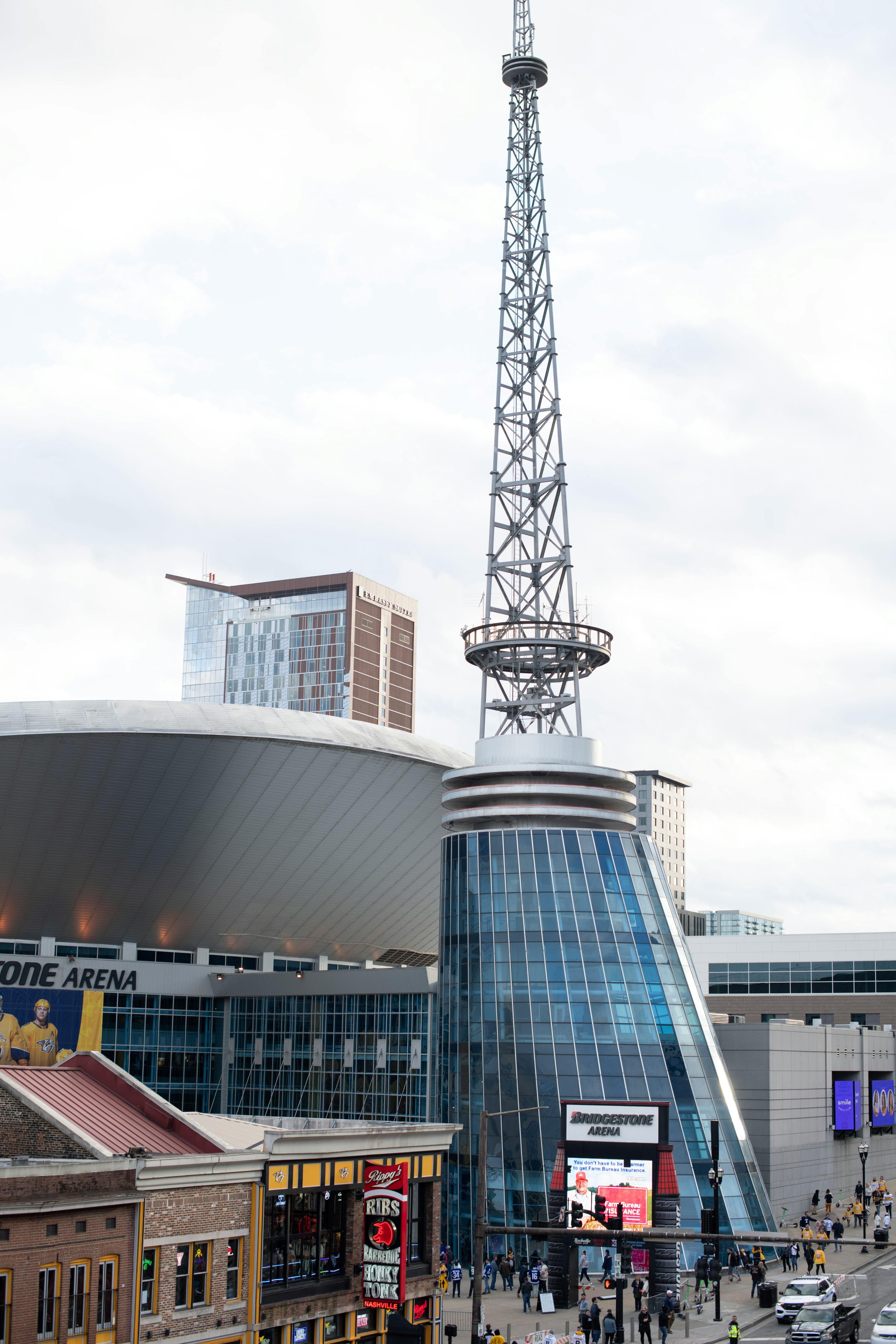A tall radio tower with a circular platform near the middle, situated next to the Bridgestone Arena in Nashville, Tennessee, with people and signs visible at street level.