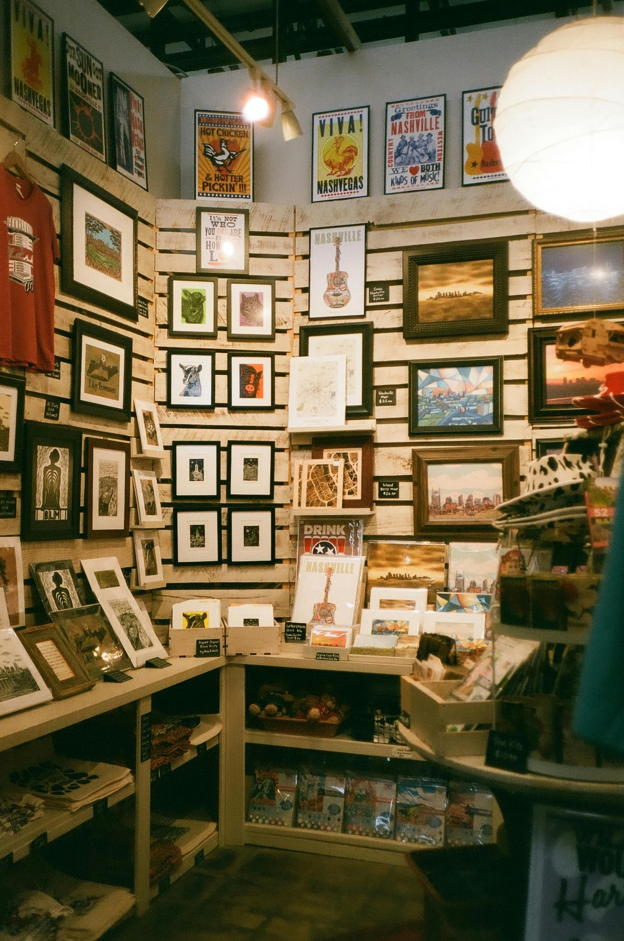 A wall filled with framed art and posters, displaying Nashville-themed artwork, vintage cityscapes, and decorative images, with shelves holding calendars, signs, and small souvenirs.