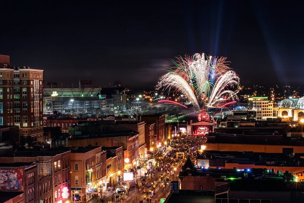 Nighttime city street with lit buildings and a crowd, fireworks exploding above with colorful lights in the sky.