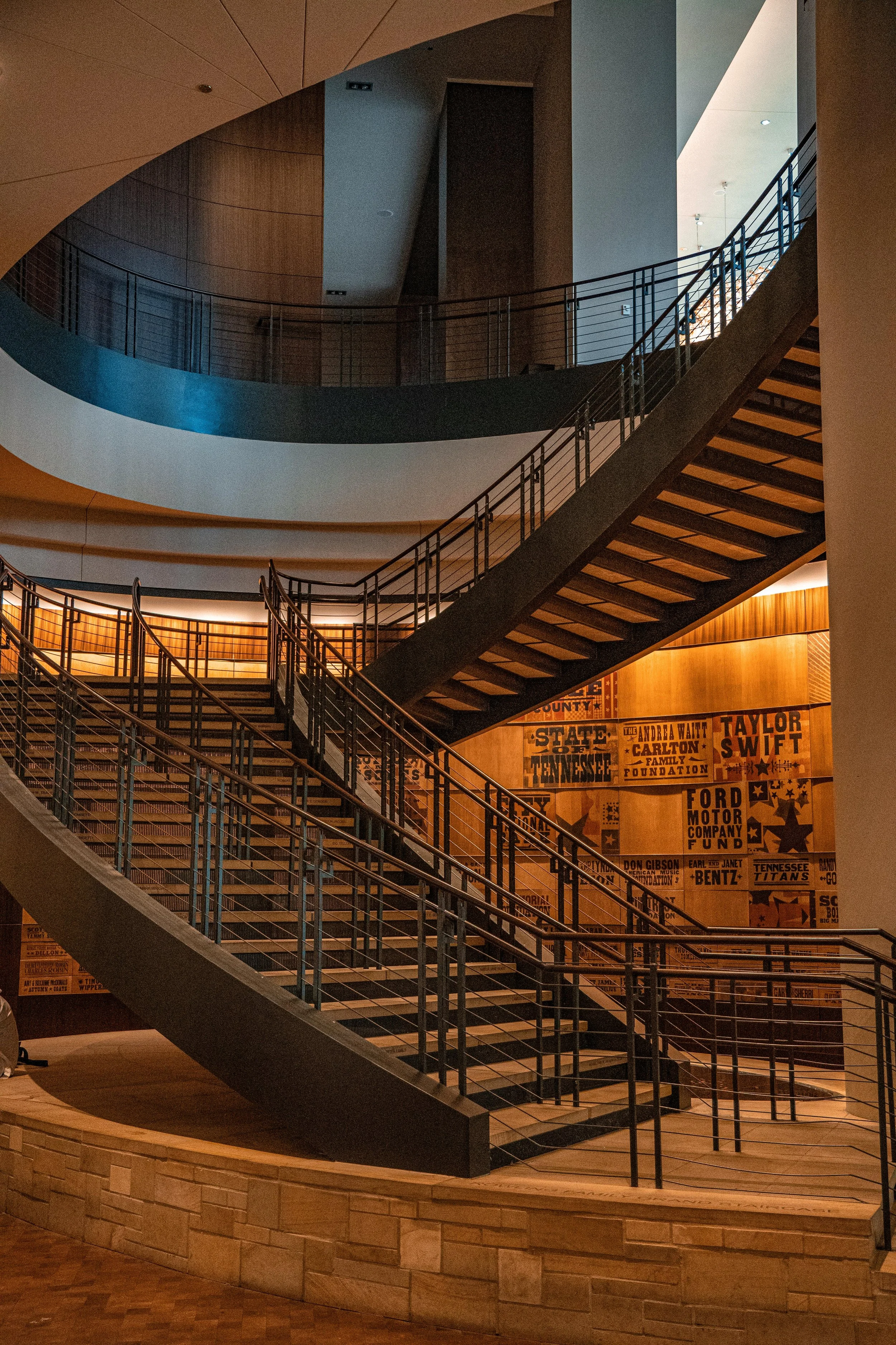 Interior of a modern building featuring a staircase with metal railings, a curved balcony, and a wooden wall with vintage posters and signs.
