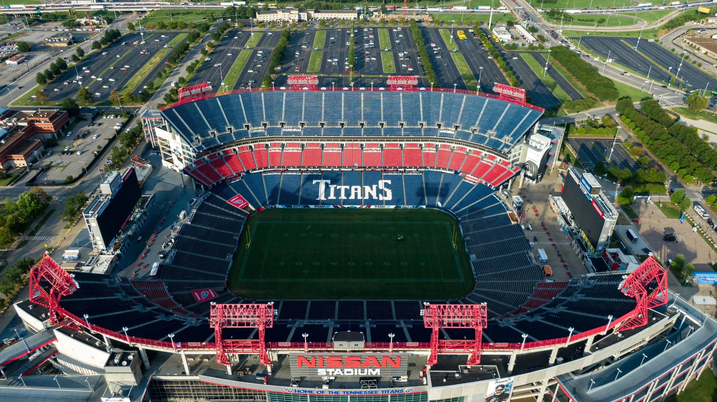 Aerial view of Nissan Stadium in Nashville, Tennessee, with a football field in the center, surrounded by red, blue, and gray seating sections, and parking lots behind the stadium.