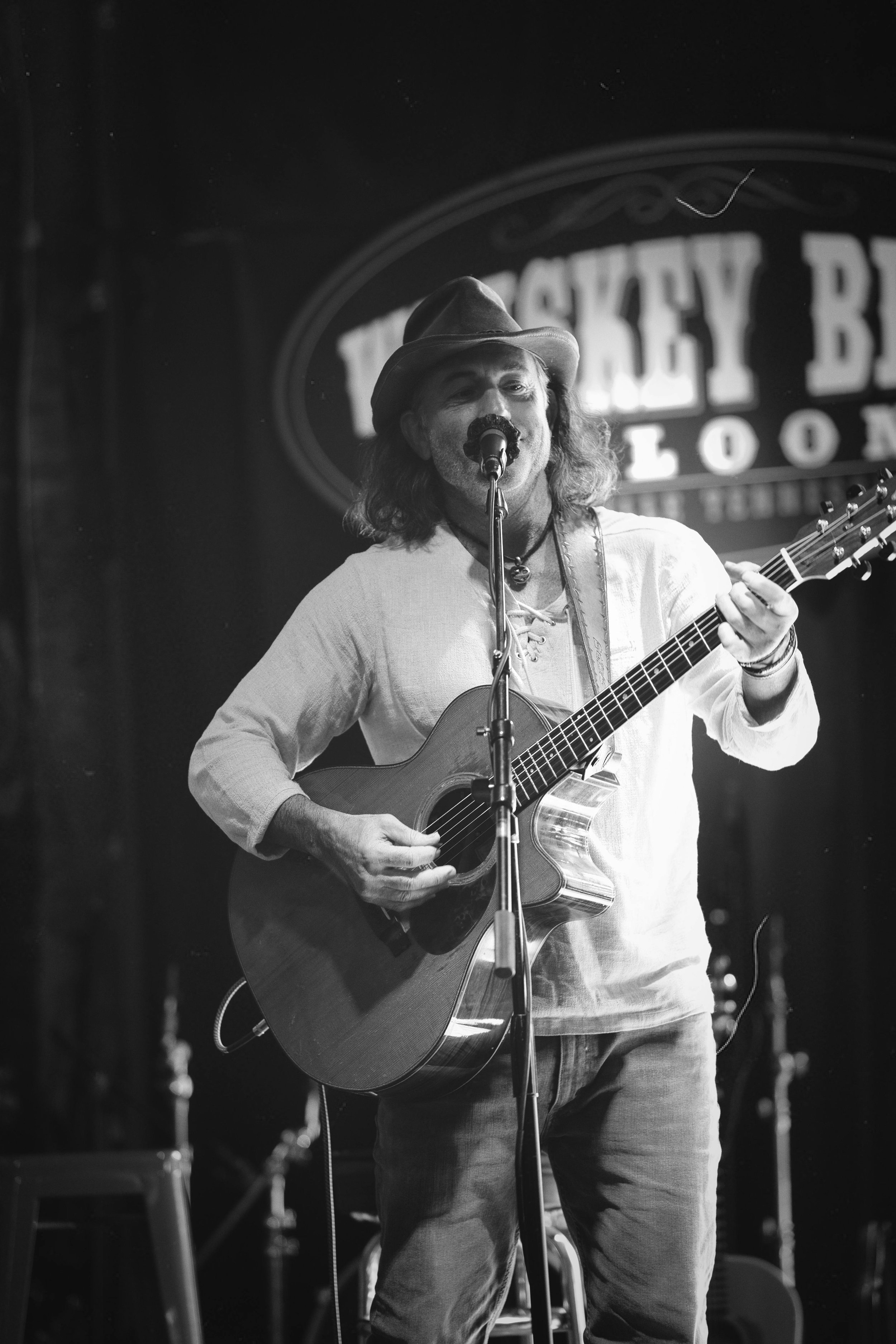 A man with long hair wearing a hat and white shirt playing an acoustic guitar and singing into a microphone on stage at a music venue.