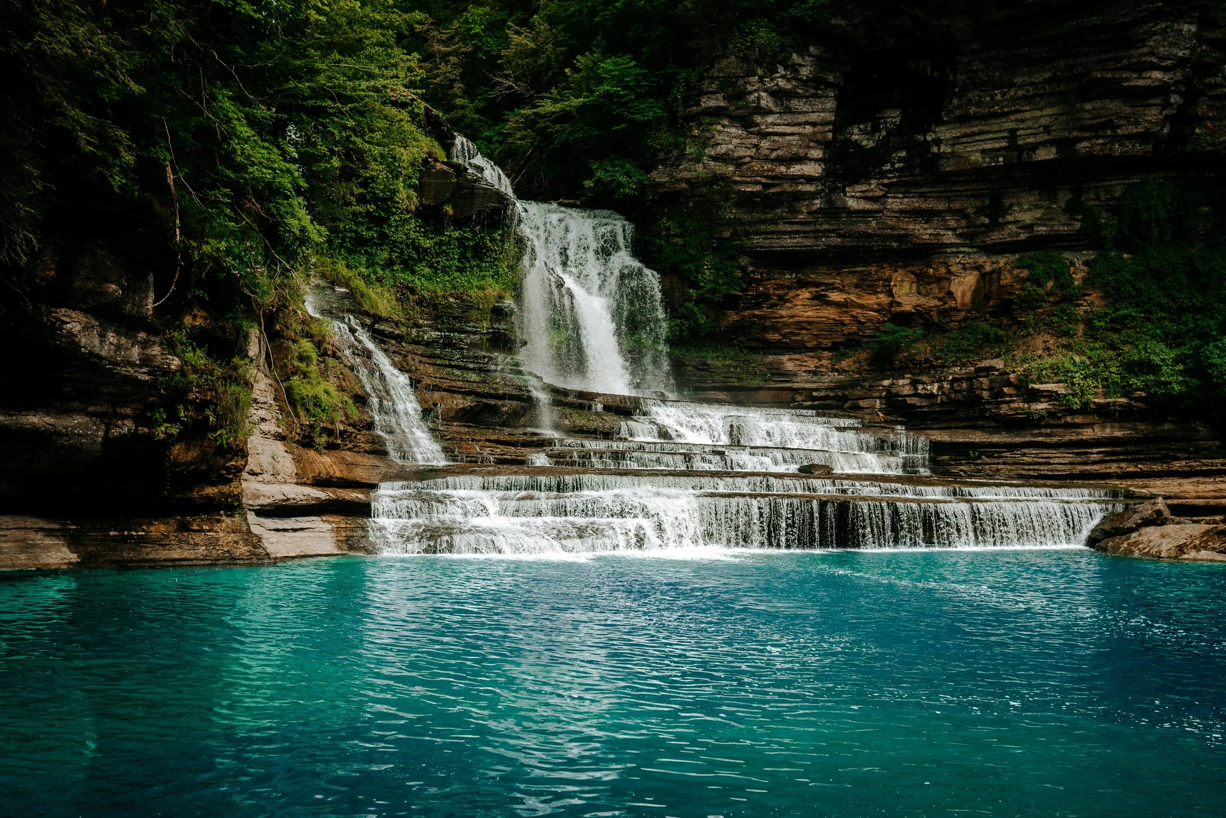 A turquoise waterfall cascading over rocks and green foliage in a natural outdoor setting.