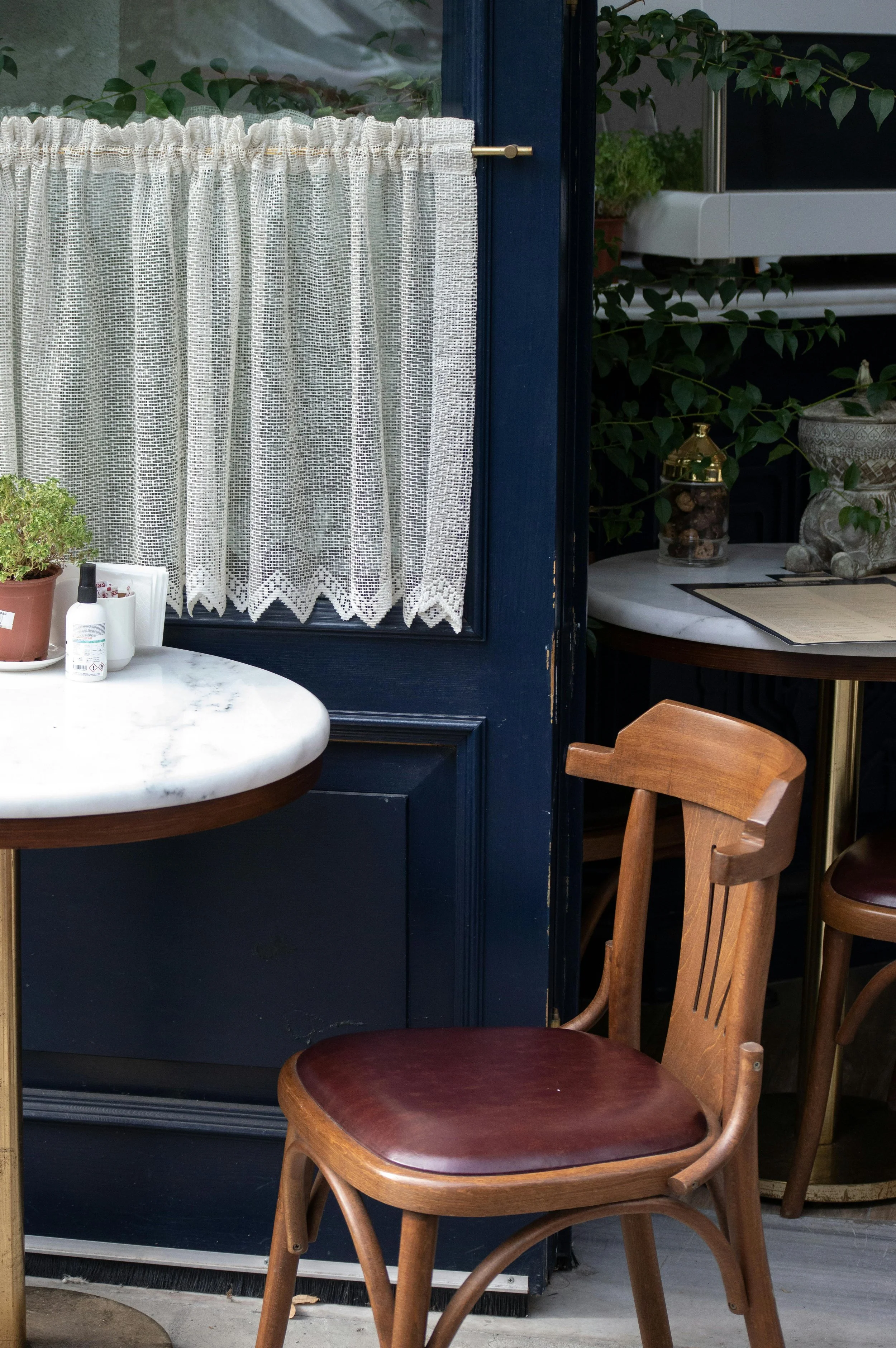 Cozy cafe setting with a wooden chair and marble table, decorated with a plant and small bottle, next to a blue door with lacy curtains.