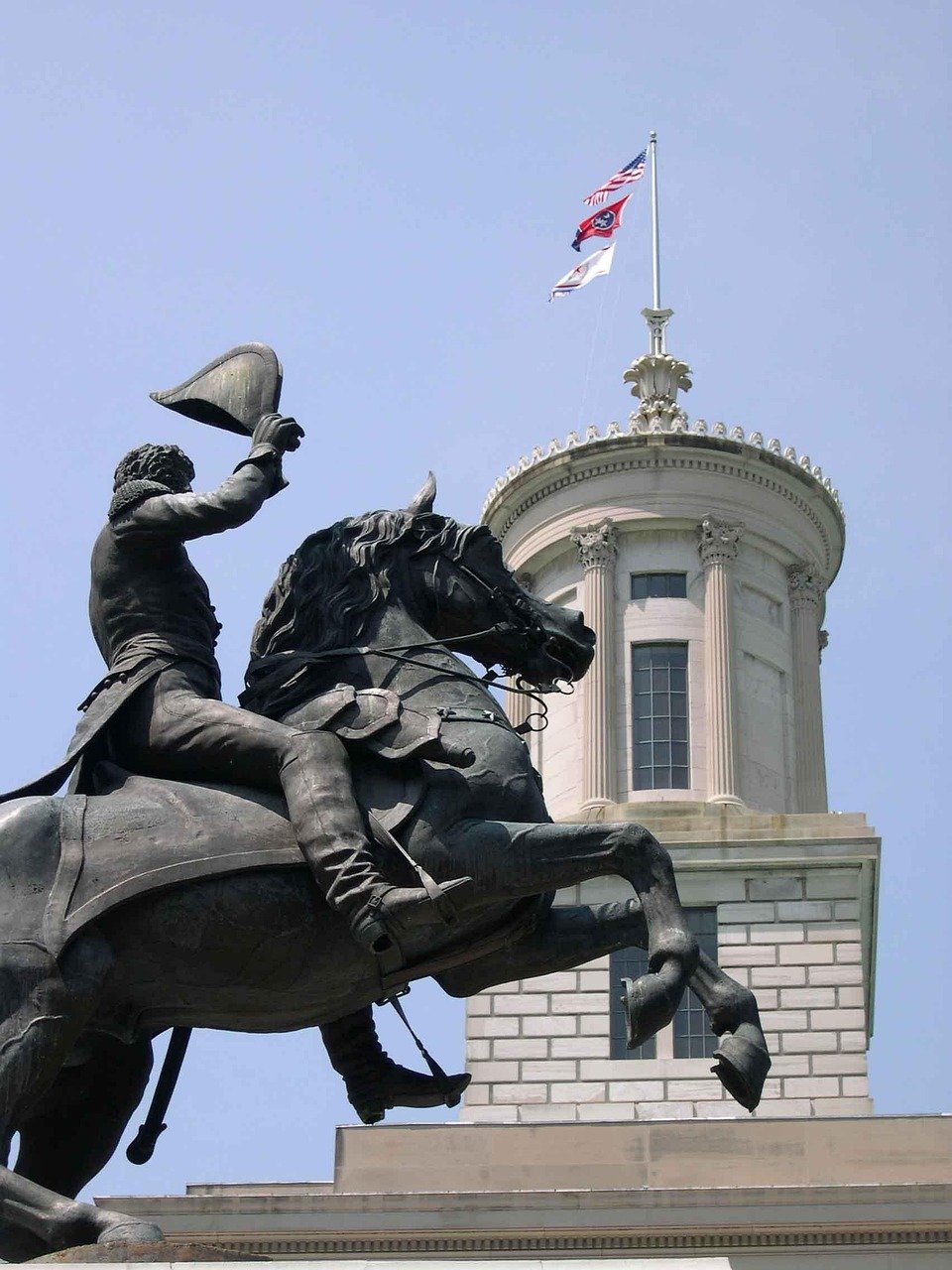 A bronze statue of a man on horseback holding a hat in the air in front of a white historic building with flags on top, including an American flag.