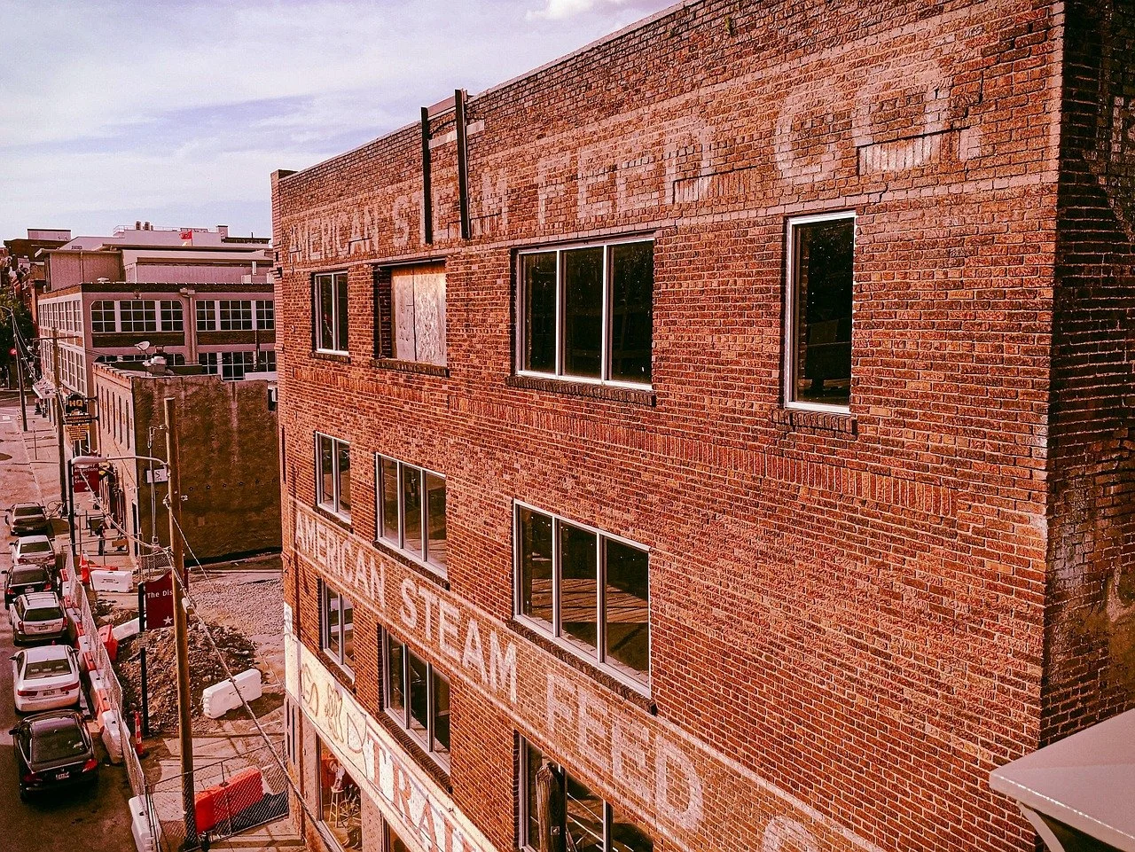 An old brick building with large windows, some with broken or boarded-up panes, and weathered faded signage for American Steam Food.