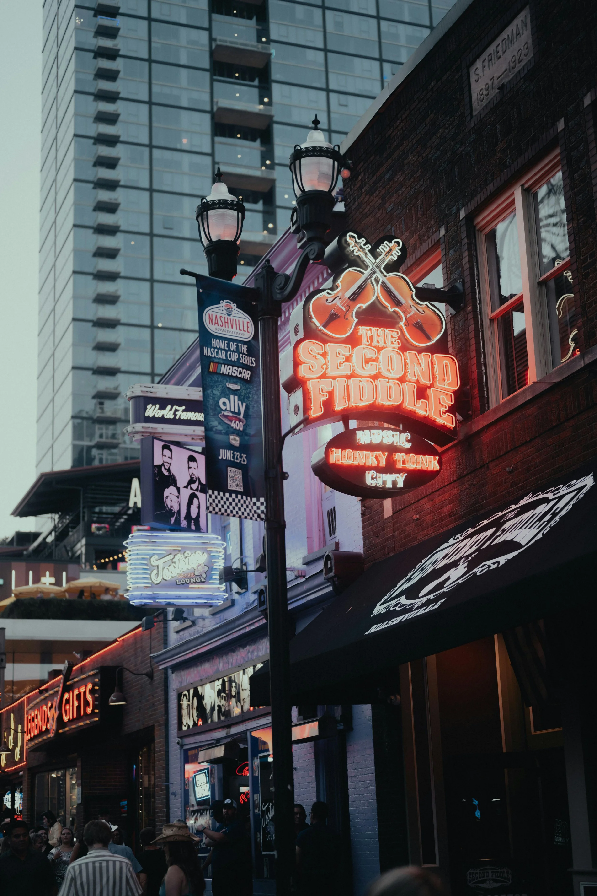 Neon sign for the Second Fiddle music venue with crossed guitars, in an urban area with people, storefronts, and tall buildings in downtown Nashville.