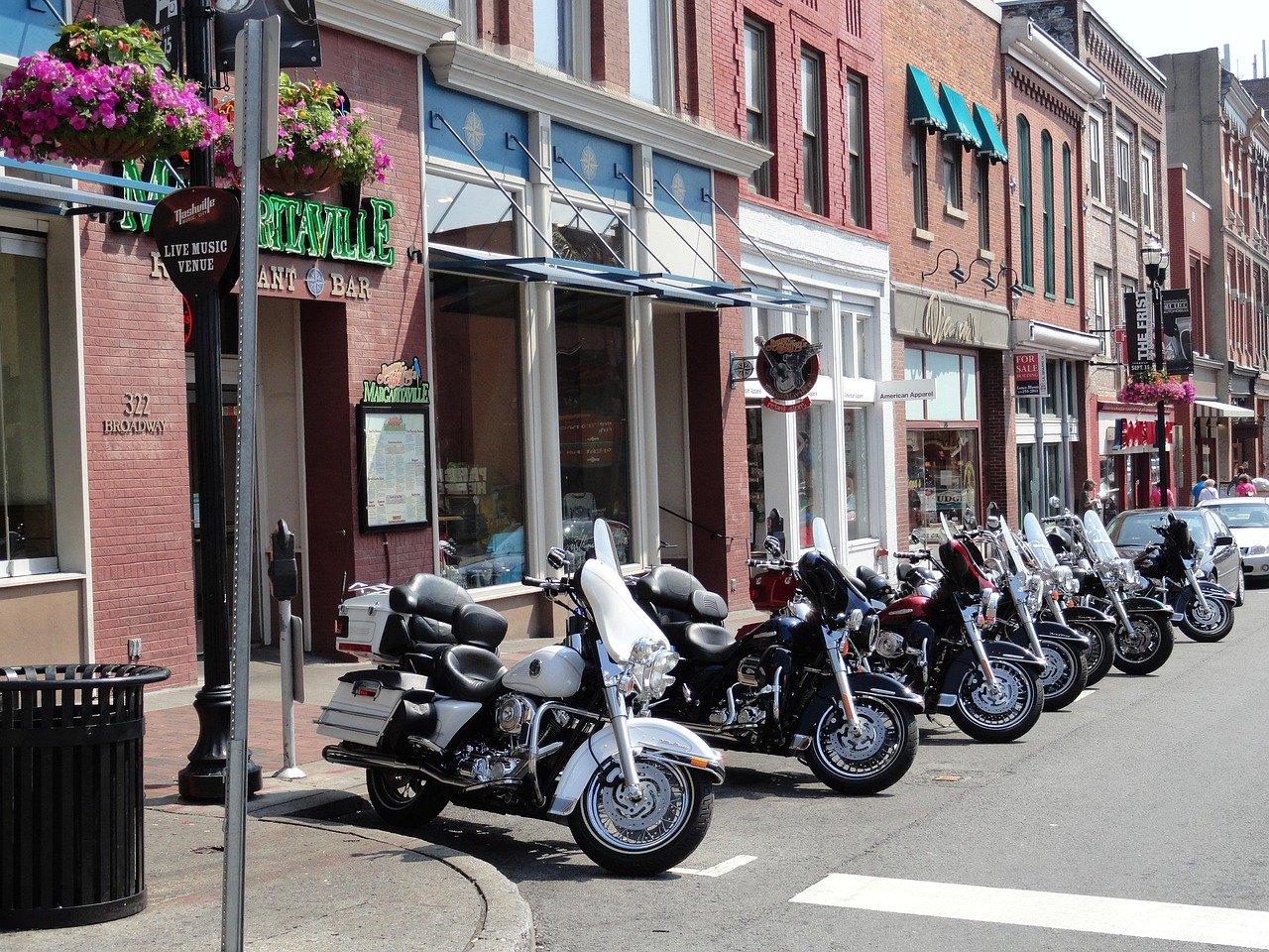 Row of parked motorcycles on a city street with storefronts, hanging flower baskets, and pedestrians walking in the background.