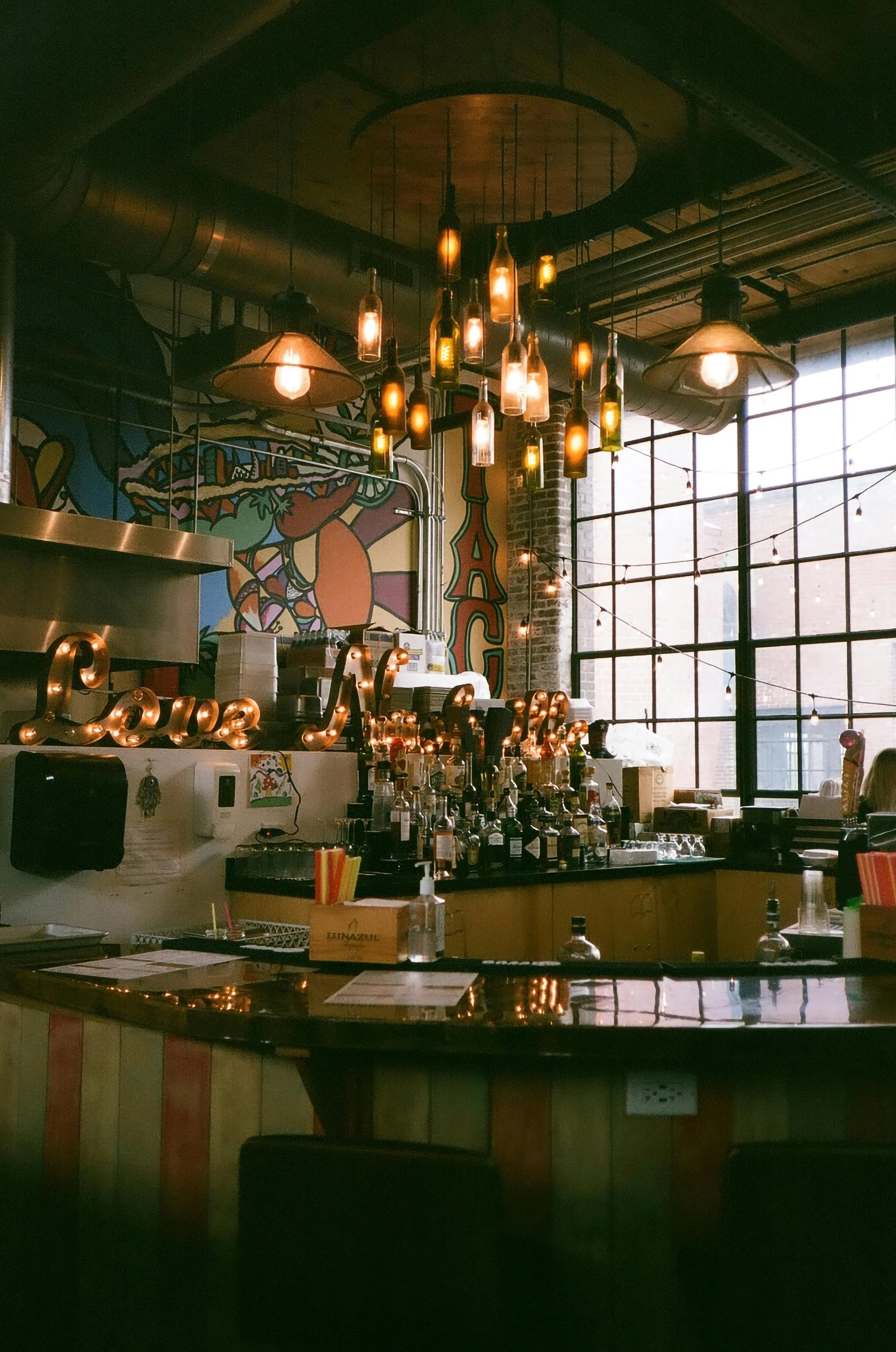 Indoor view of a bar with warm lighting, bottles on the counter, a neon sign, artistic wall mural, and large industrial windows.