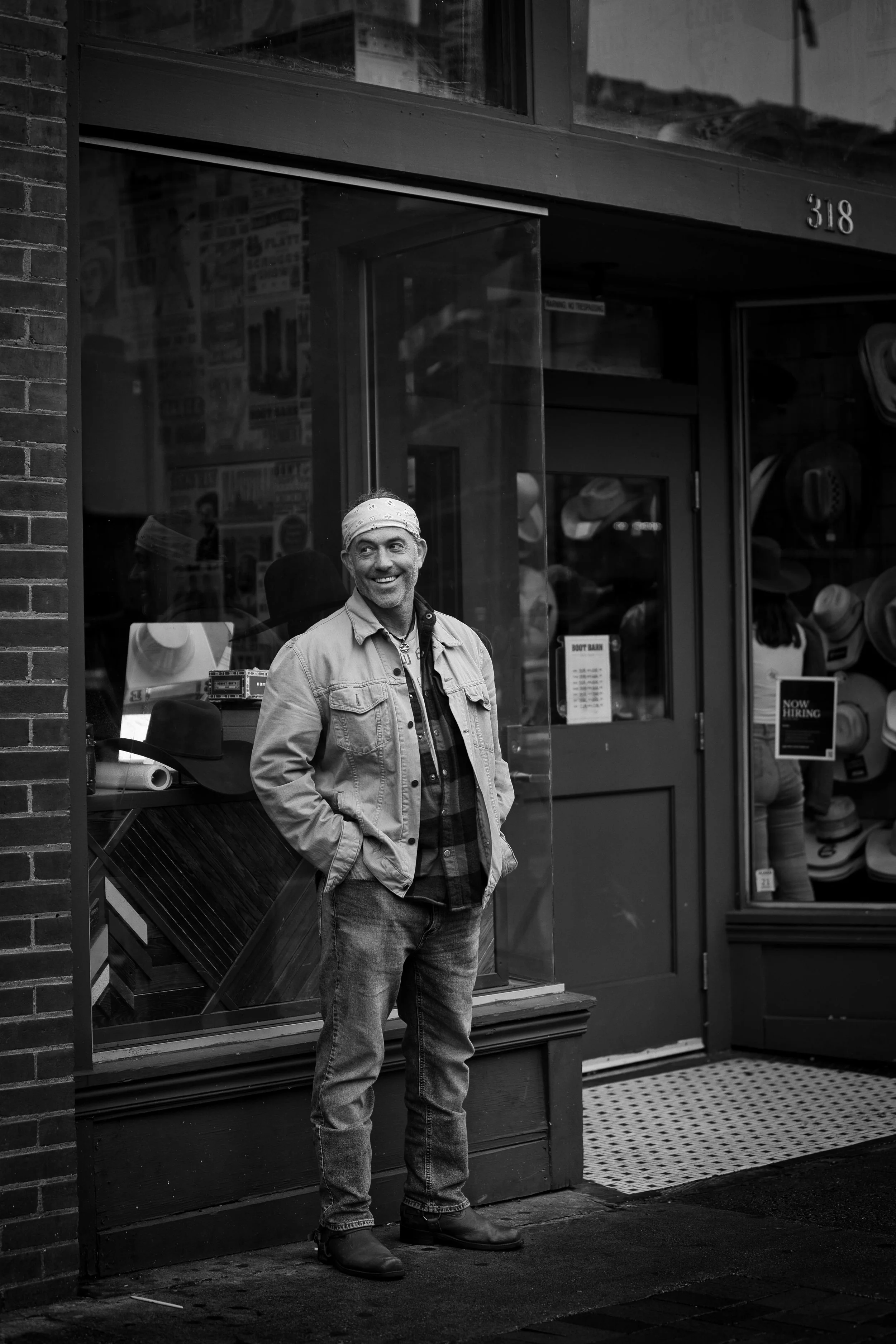 A man standing outside a shop window with a smile, looking to the side. The shop has various hats displayed inside, and signs indicating it is hiring.