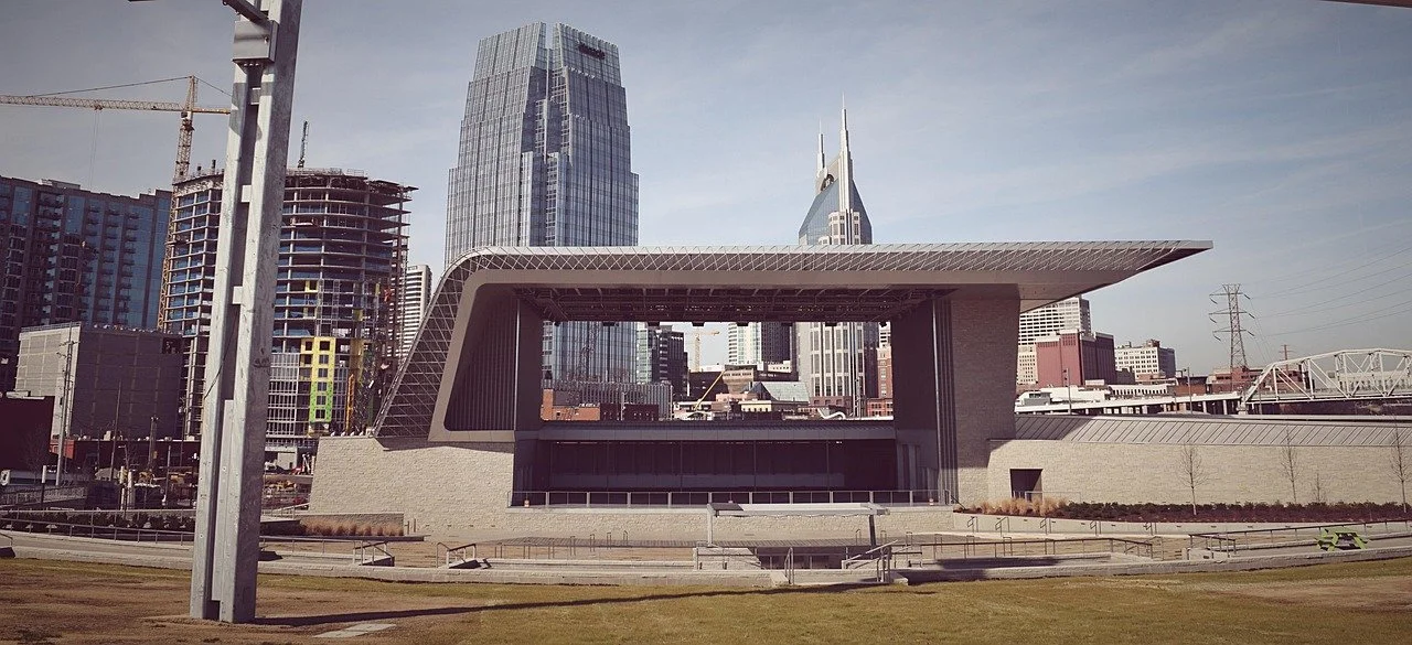 Modern cityscape with tall skyscrapers, including one with a unique angled roof, and an under-construction building in the background. A large, contemporary public building with a flat, overhanging roof is in the foreground, along with grassy area an