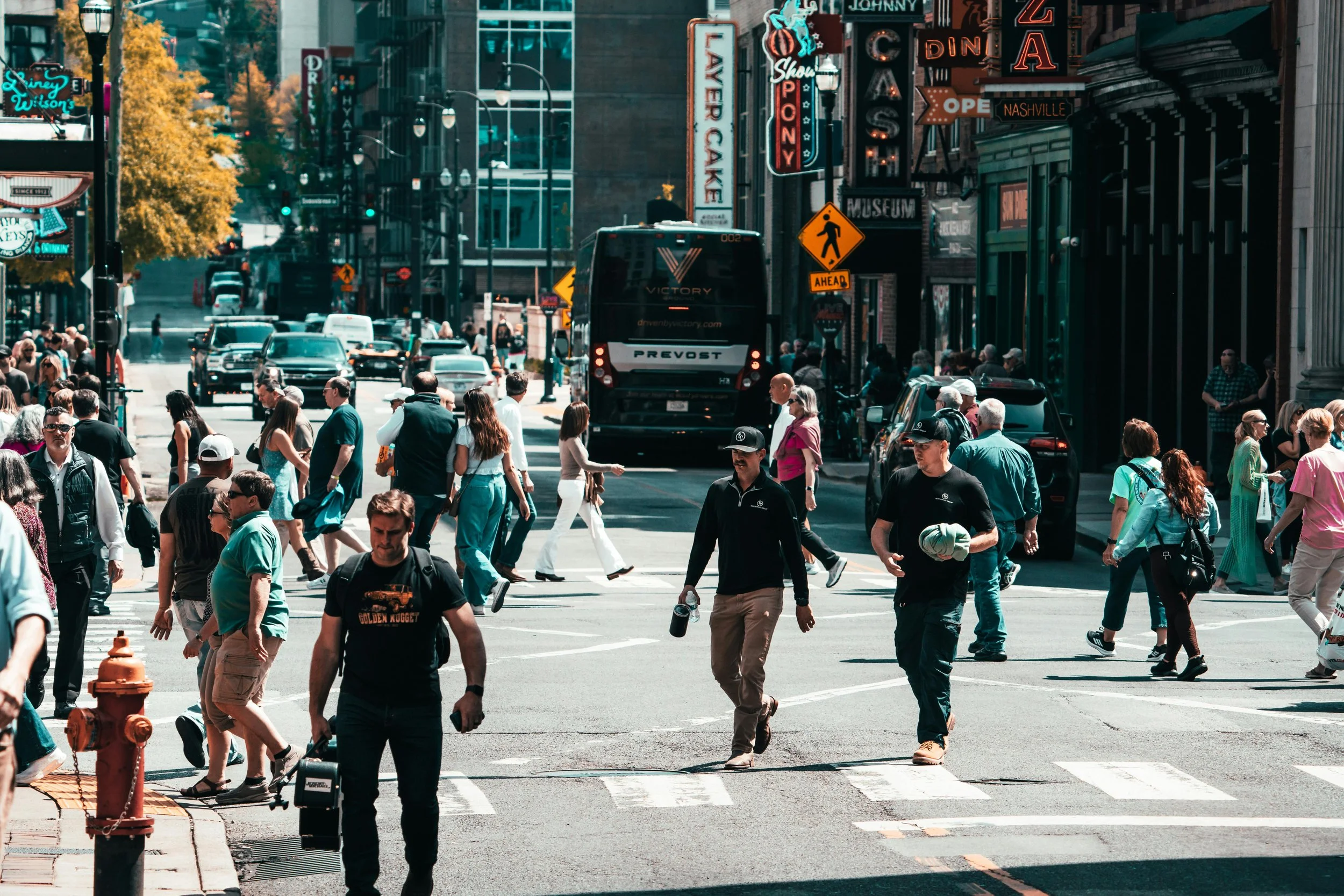 A busy city street with pedestrians crossing, cars parked and driving, and a bus in the background. Buildings with neon signs and storefronts line the street.