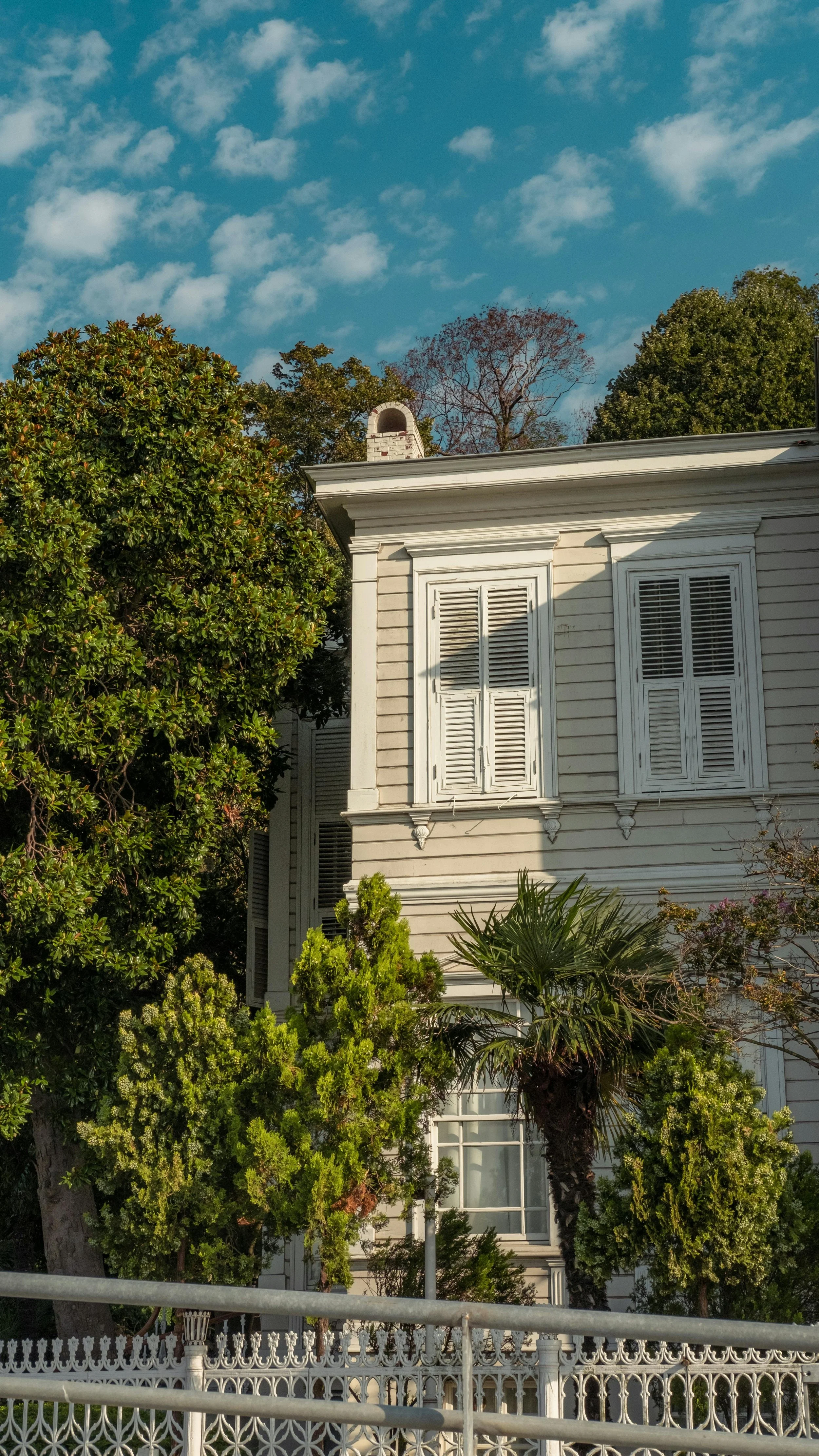 Historic wooden house with white shutters and ornate trim, surrounded by lush green trees, against a partly cloudy blue sky.