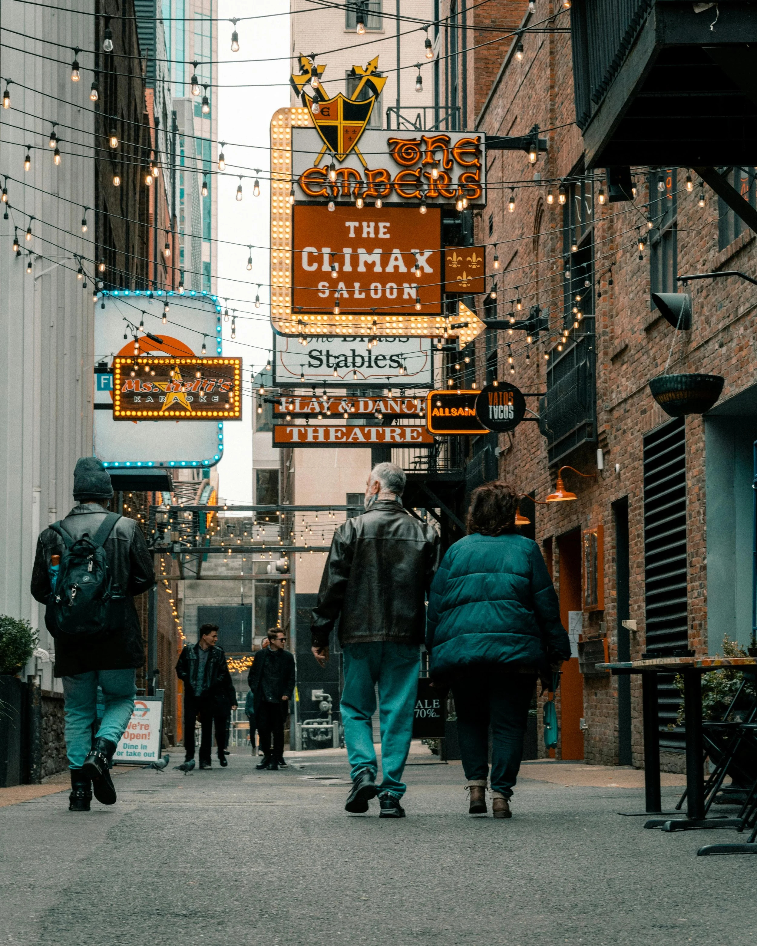 People walking down a city alleyway decorated with string lights and neon signs for bars, a casino, and a theatre, with brick and building facades on either side.