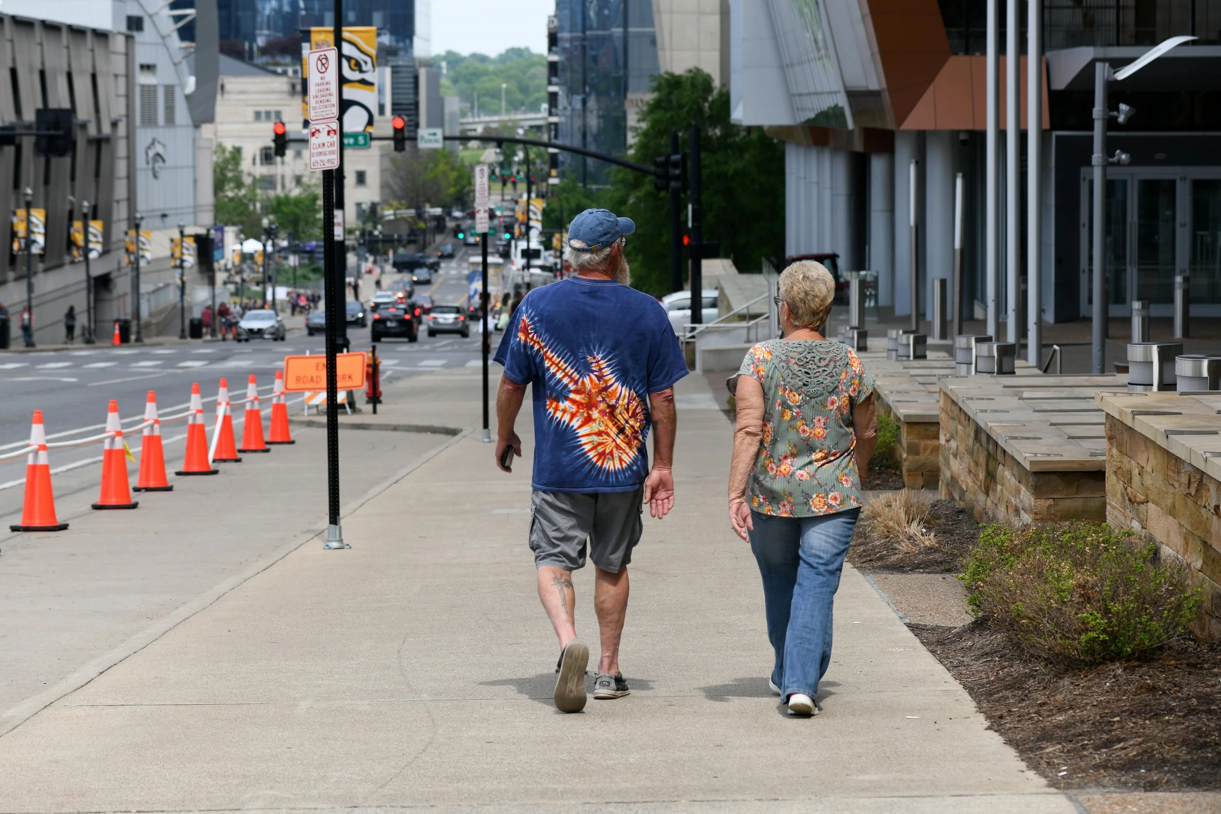 Two people walking on a city sidewalk, with one wearing a tie-dye shirt and the other in a floral top, in front of a street with cars and orange construction cones.