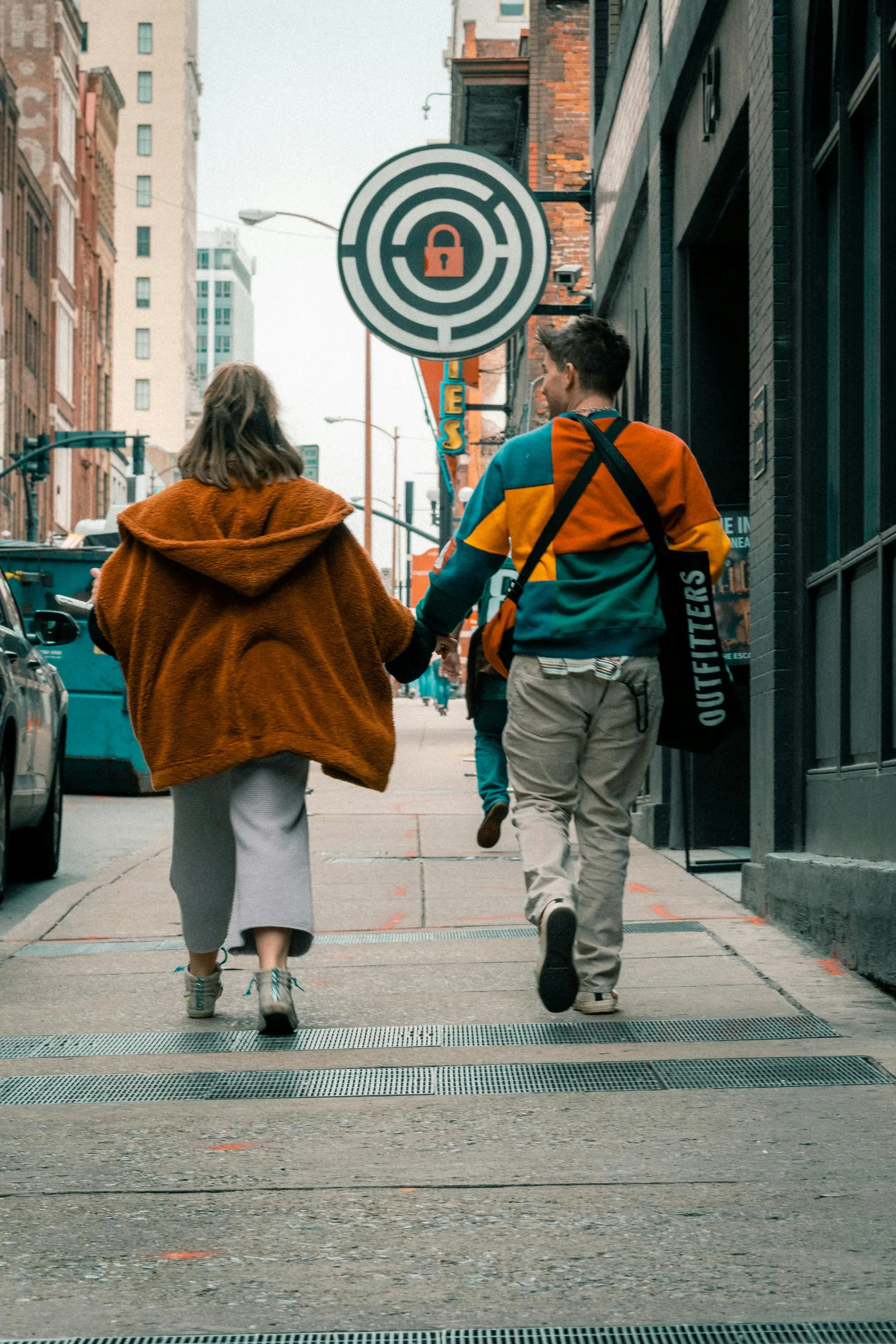 A young man and woman holding hands walk down a city sidewalk, with tall buildings and street signs visible in the background.