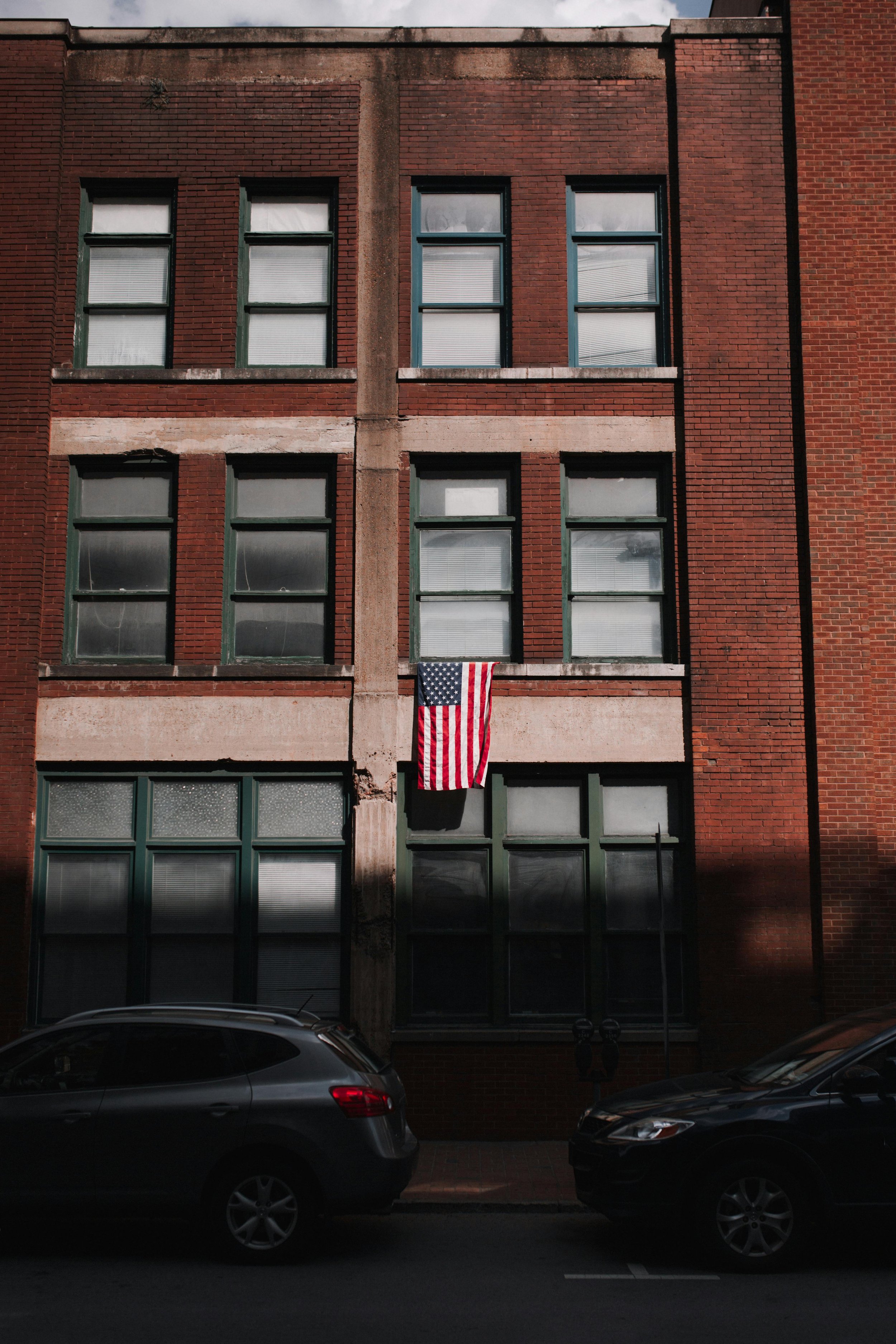 A red brick building with multiple windows and an American flag hanging in the middle, with cars parked in front.