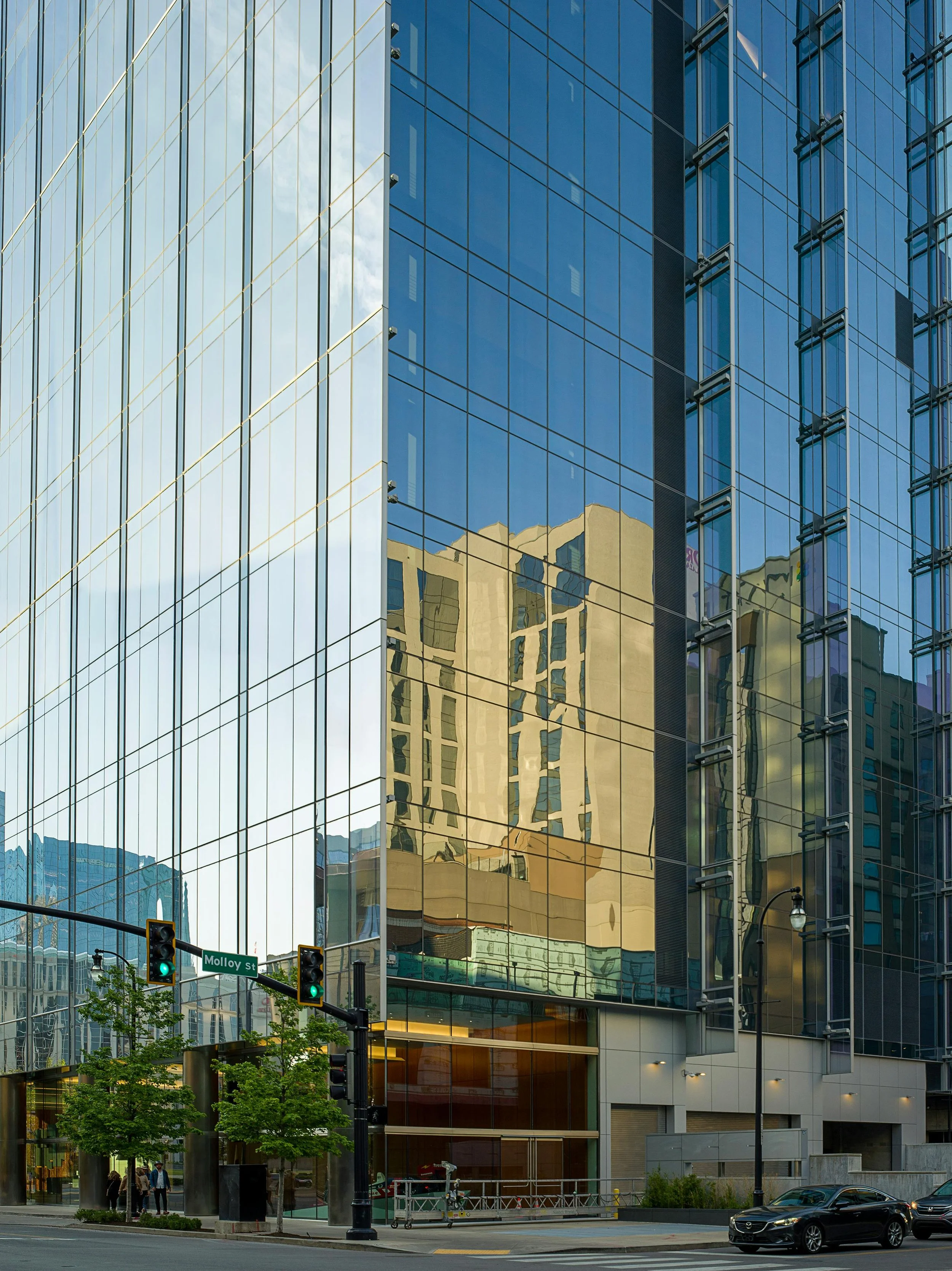 Reflections of a modern high-rise building and trees on glass building windows near an intersection with a traffic light and cars.