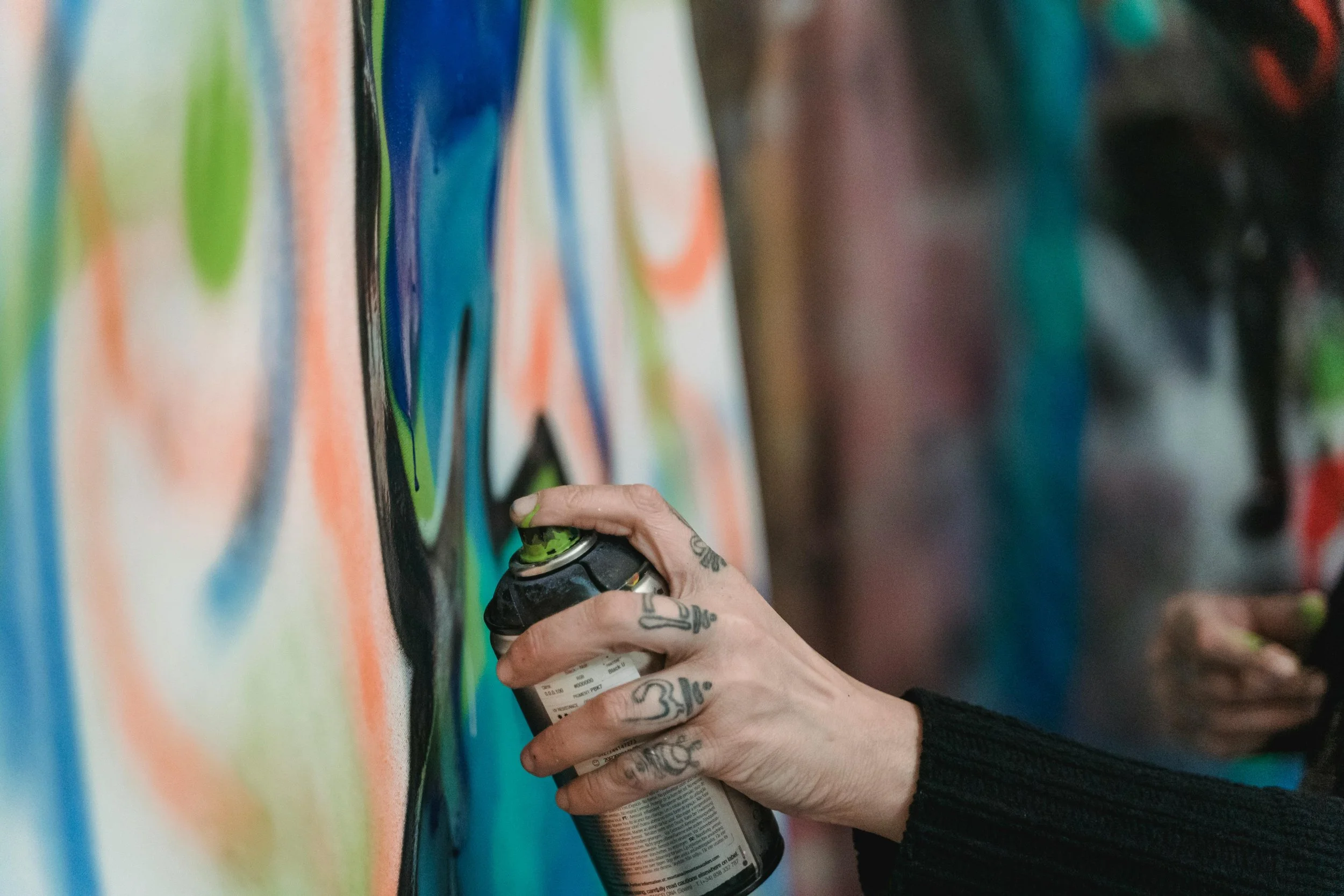 Person with tattoos on their fingers spray-painting a colorful abstract mural on a wall.