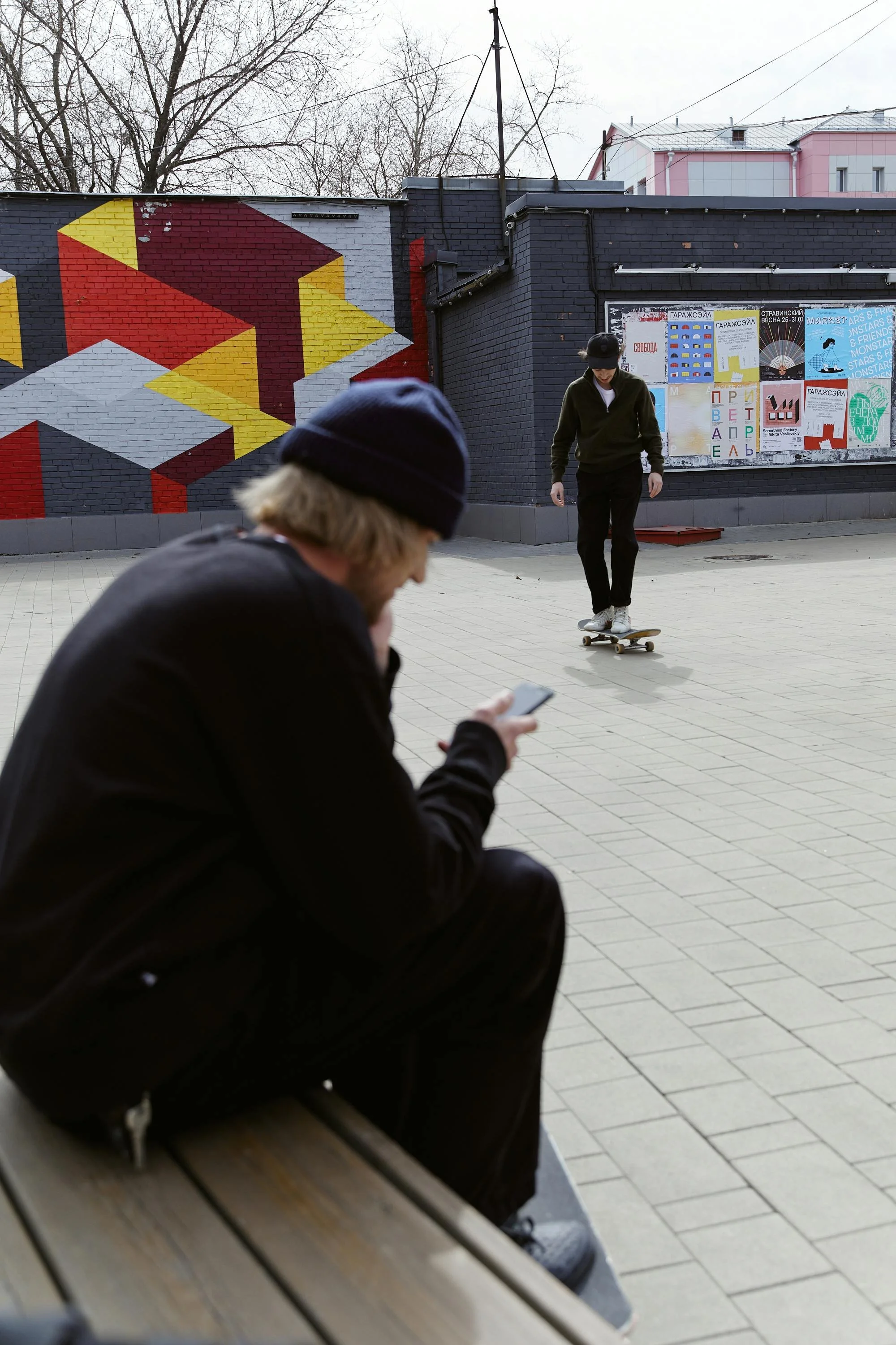 A person sitting on a bench looking at a phone in front of a brick wall with abstract mural art and a bulletin board, while a skateboarder rides nearby on paved ground.