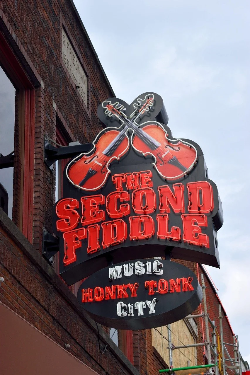 Neon sign for The Second Fiddle, a music venue in Honky Tonk City, featuring two crossed violins with a red and black color scheme, on a brick building