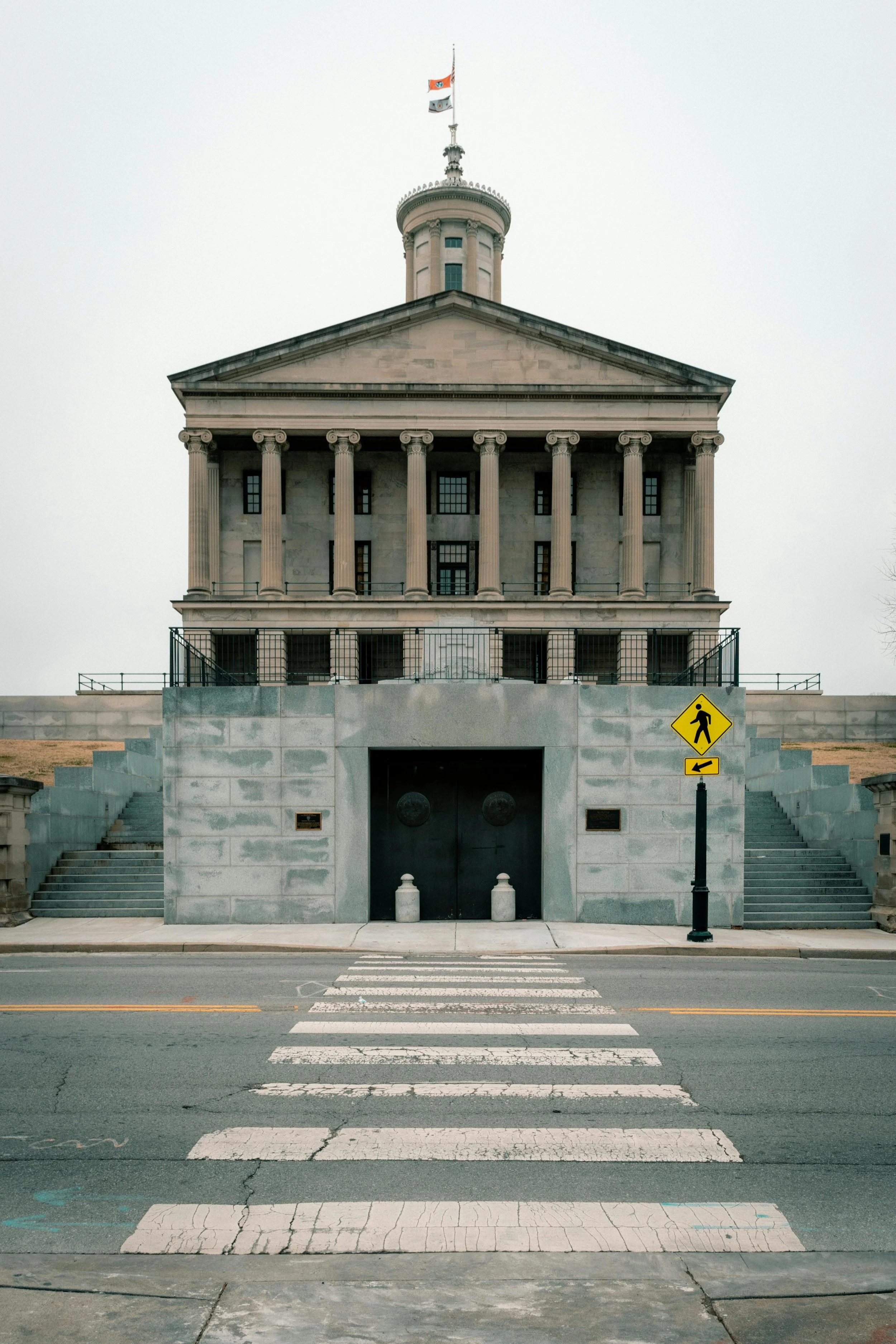 Historic courthouse with Greek Revival architecture, featuring tall columns and a central tower, viewed from a street with a pedestrian crosswalk and a yellow traffic sign.