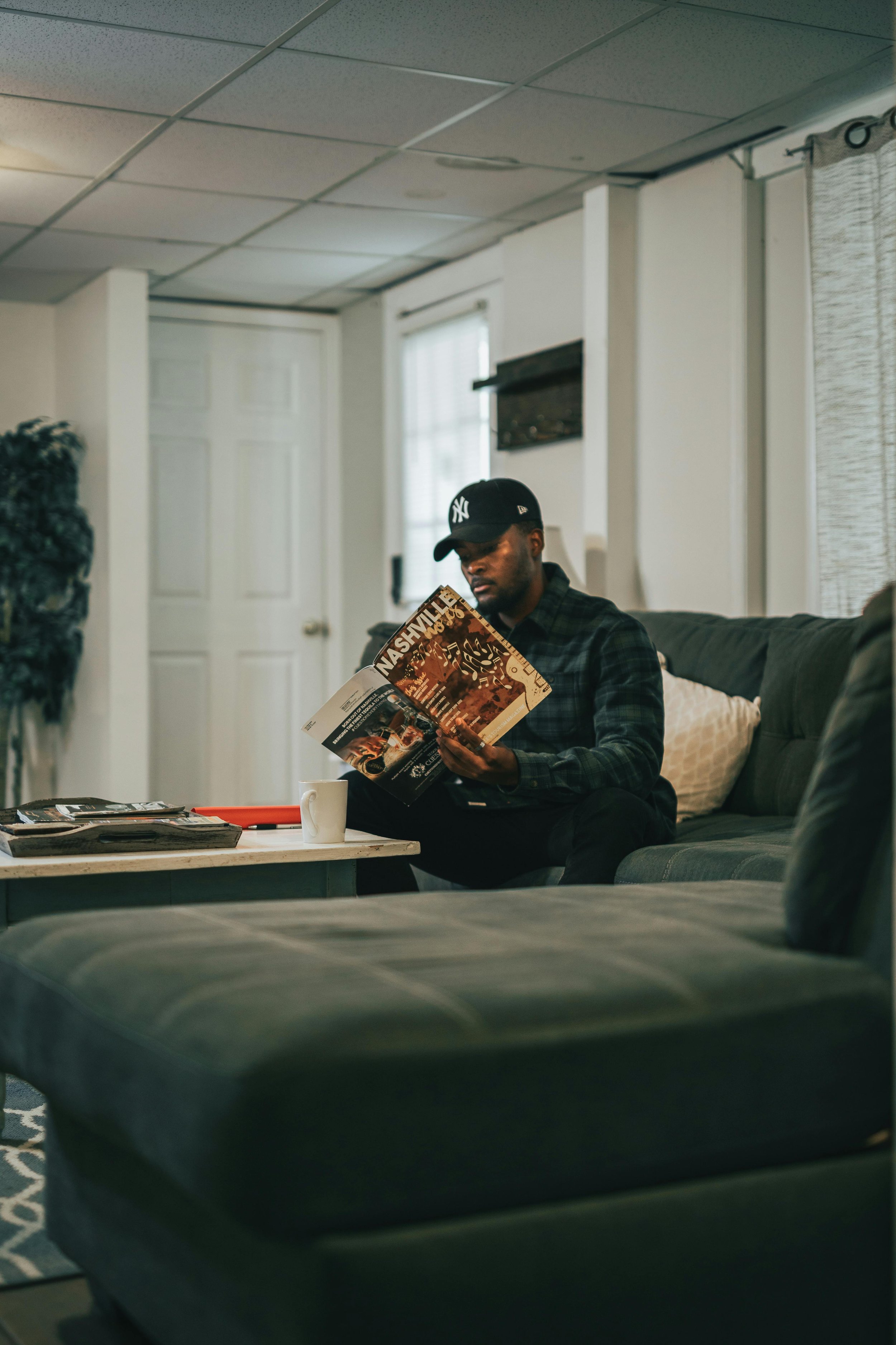A man wearing a black baseball cap and checkered shirt sitting on a dark green couch, reading a magazine titled 'Nashville' in a living room with a white door, windows, and a coffee table with magazines and a mug.