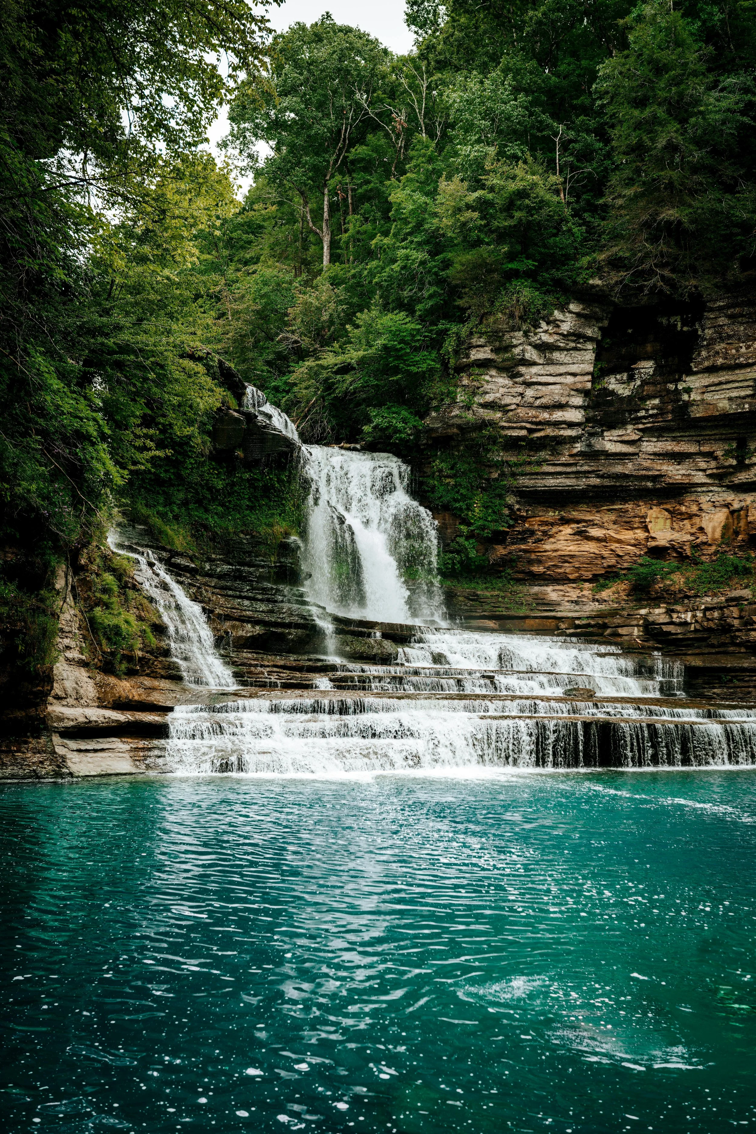 A multi-tiered waterfall flowing into a turquoise pool surrounded by green trees and rocky cliffs.