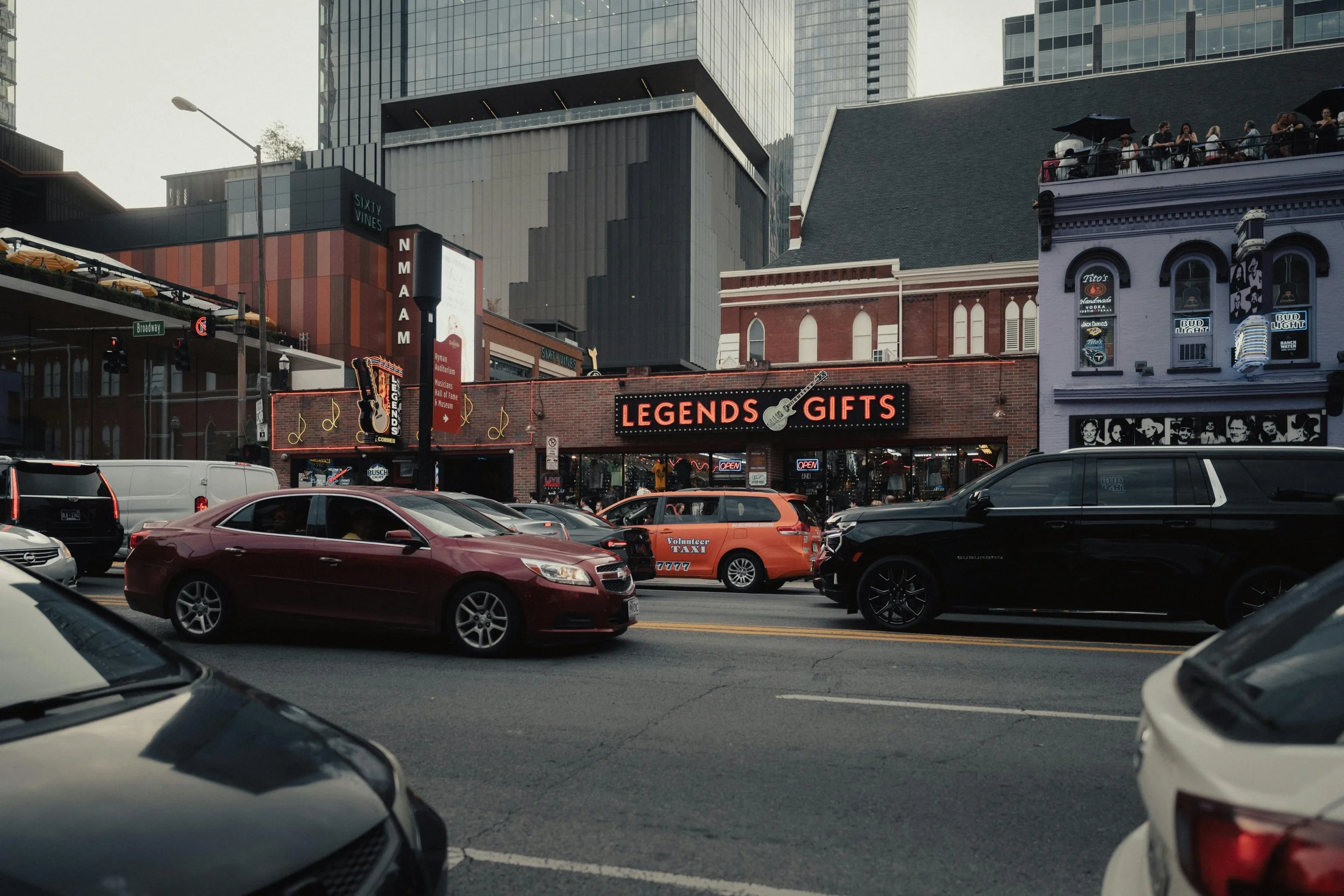 City street view with parked cars in front of a store called Legends Gifts, surrounded by tall buildings and a crowd on a rooftop terrace.